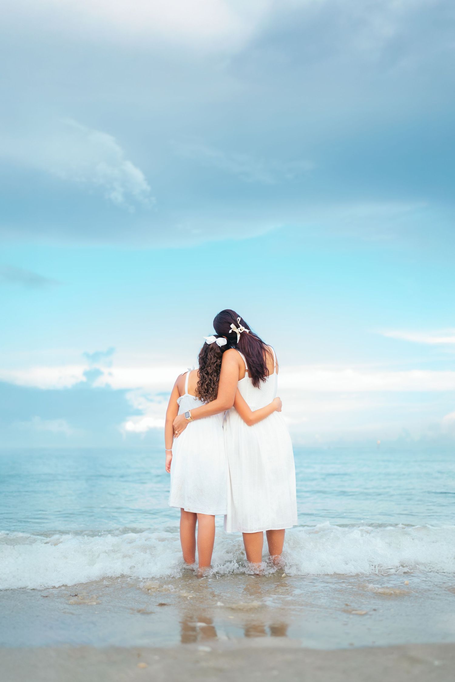 Two people in white dresses stand together at the ocean's edge during a peaceful beach sunset.