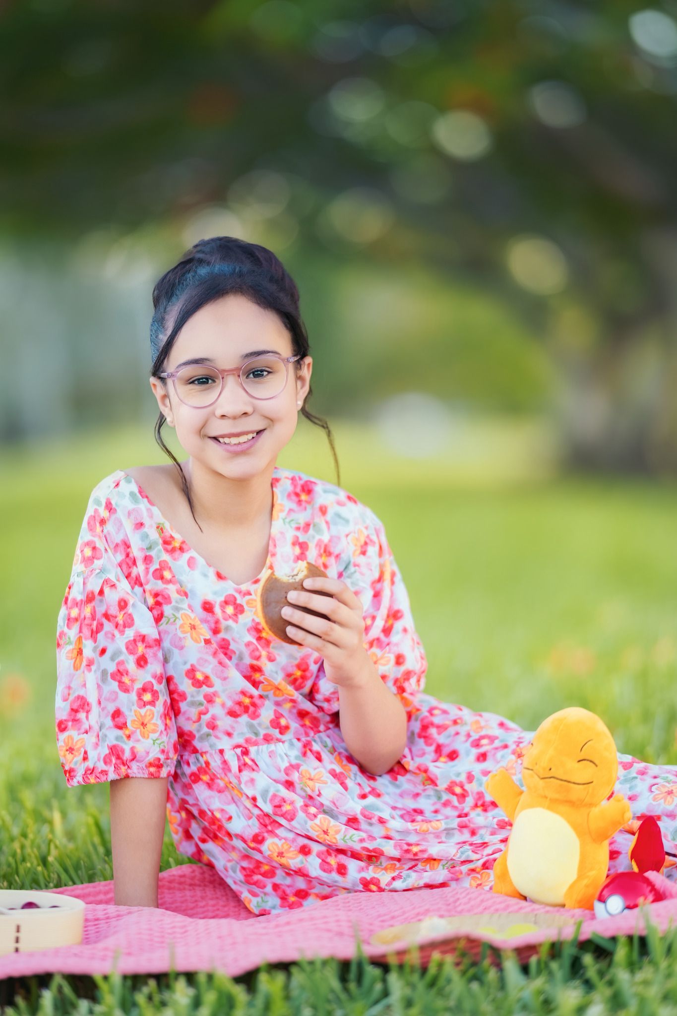 A person in a pink floral dress relaxes on a blanket outdoors with plush toys and a mobile phone.