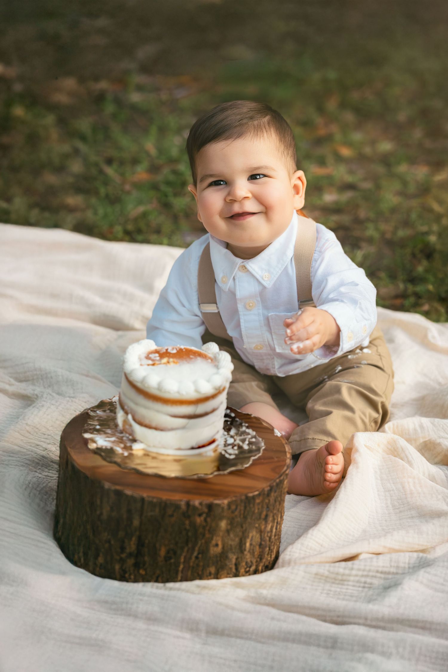 Smiling baby boy in suspenders sits on blanket outdoors beside a rustic smash cake on a wood stump.