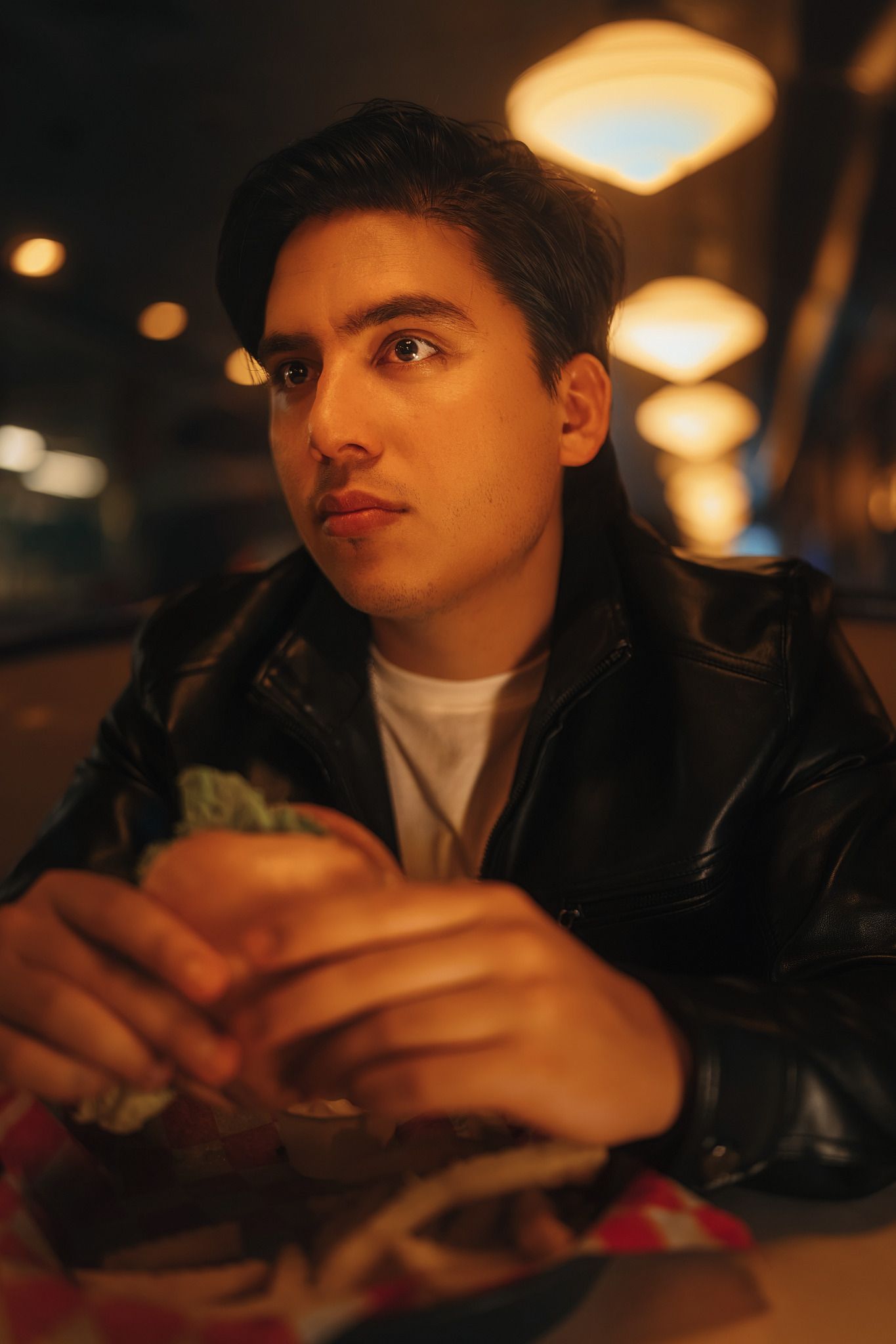 Young man in black leather jacket eating a burger at a dimly lit restaurant at night.