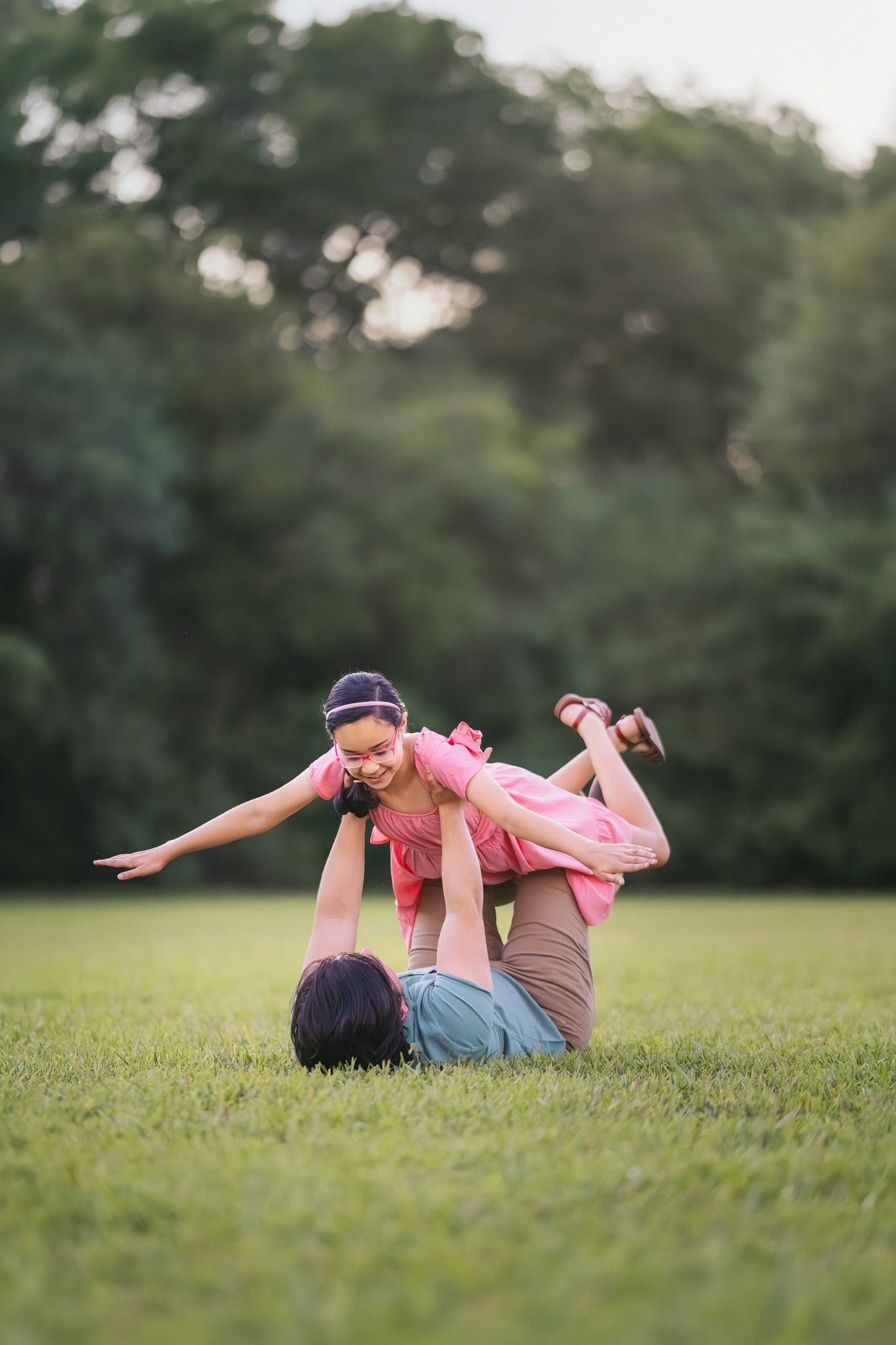 A mother lies on grass lifting her smiling daughter in a pink dress overhead in a sunny park.