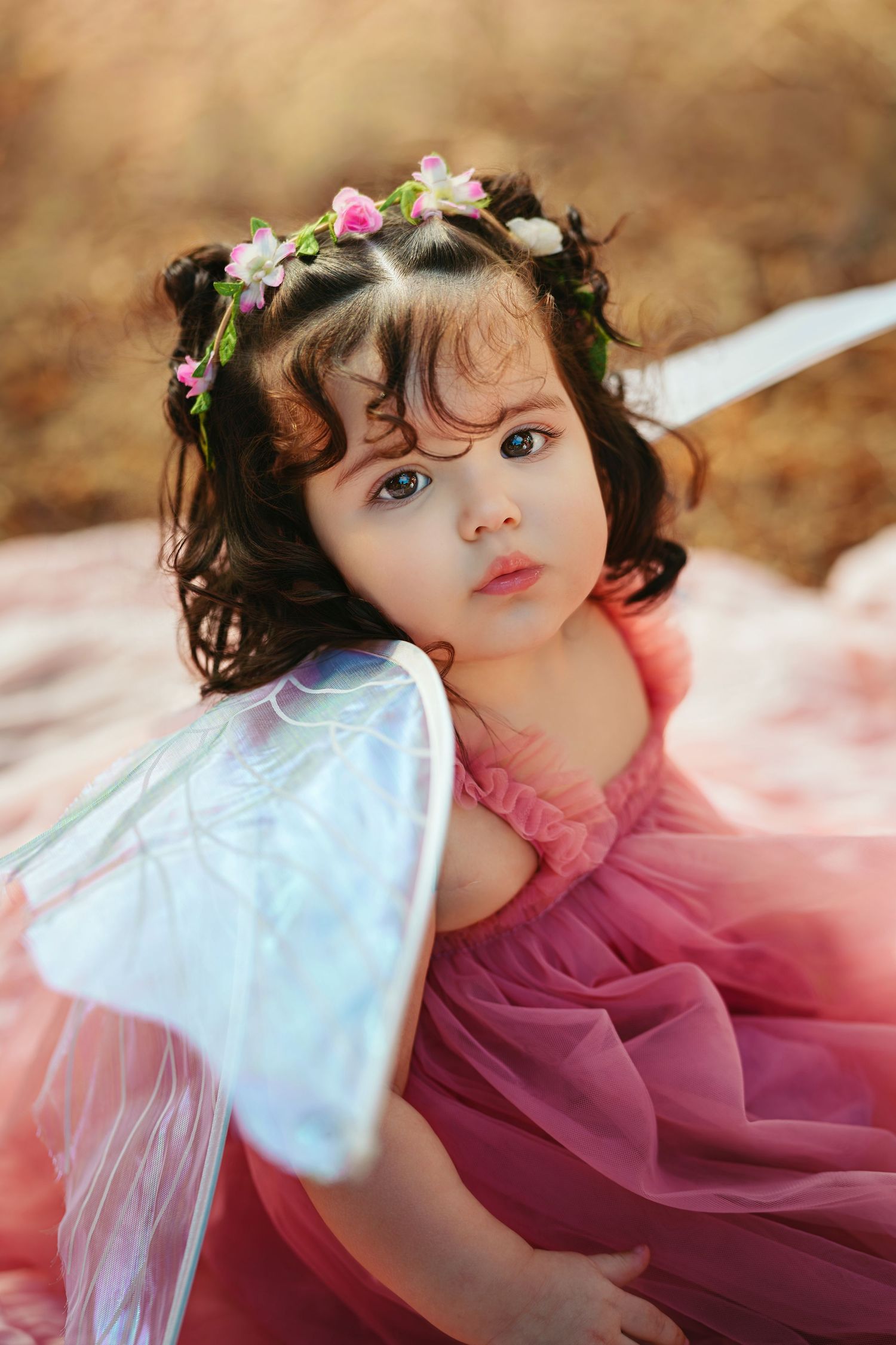 A toddler girl in a pink tulle dress with fairy wings and a floral crown poses outdoors in a dreamy portrait.