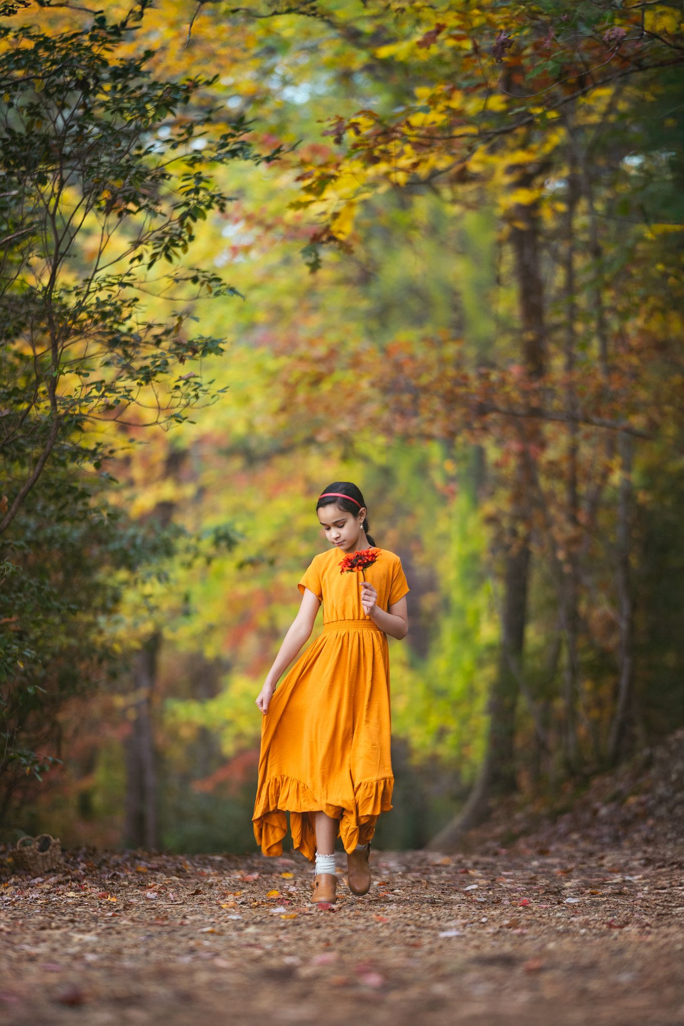 Young girl in orange dress walking on autumn forest path surrounded by colorful fall foliage.