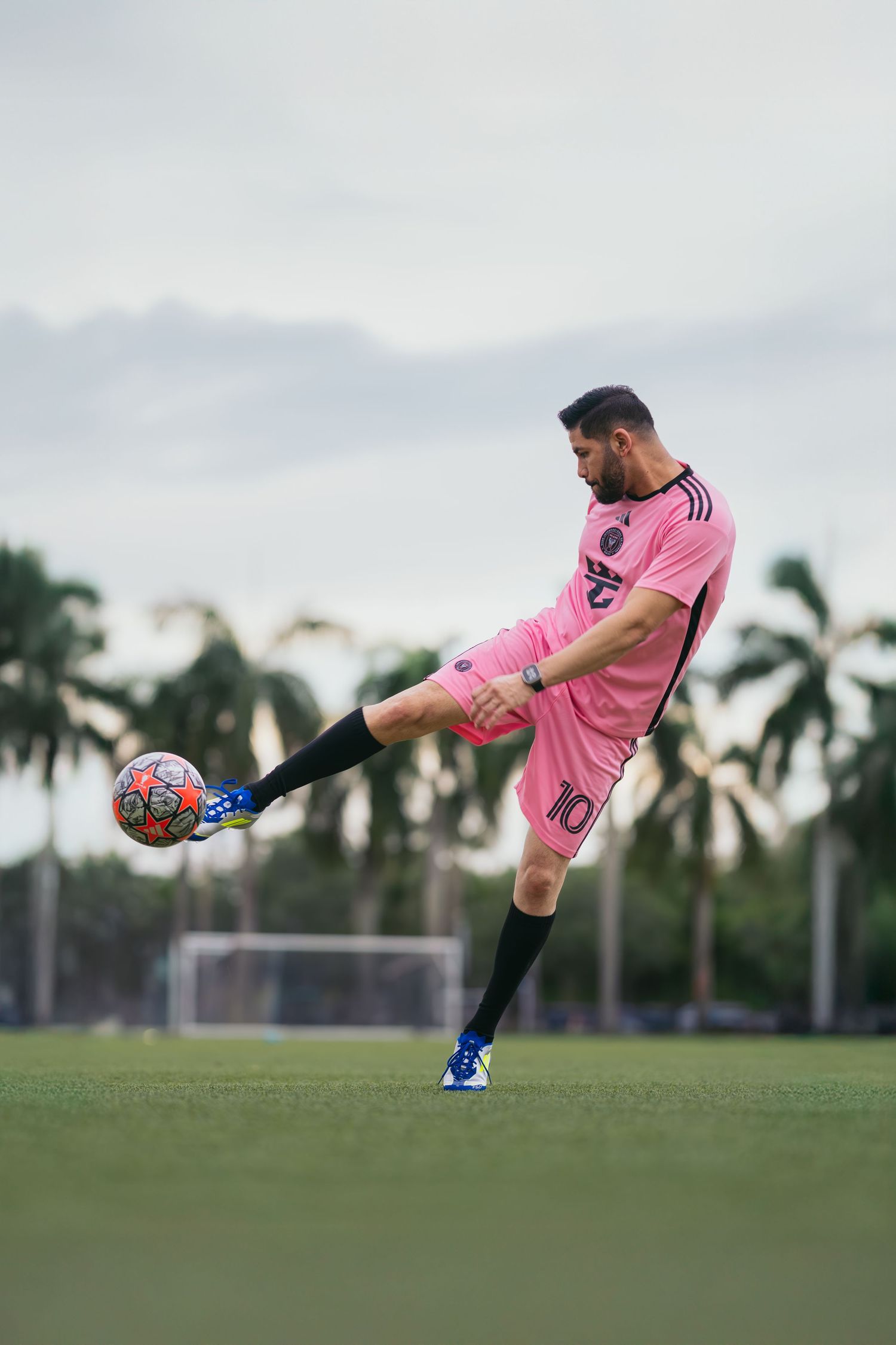 Soccer player in pink jersey number 10 kicking ball on green field with palm trees in background.