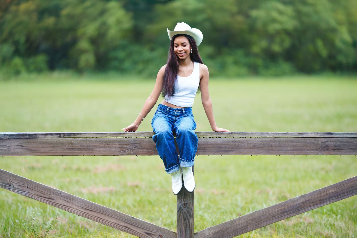 Young woman in white cowboy hat, tank top, jeans, and boots sitting on a wooden fence in a green field.