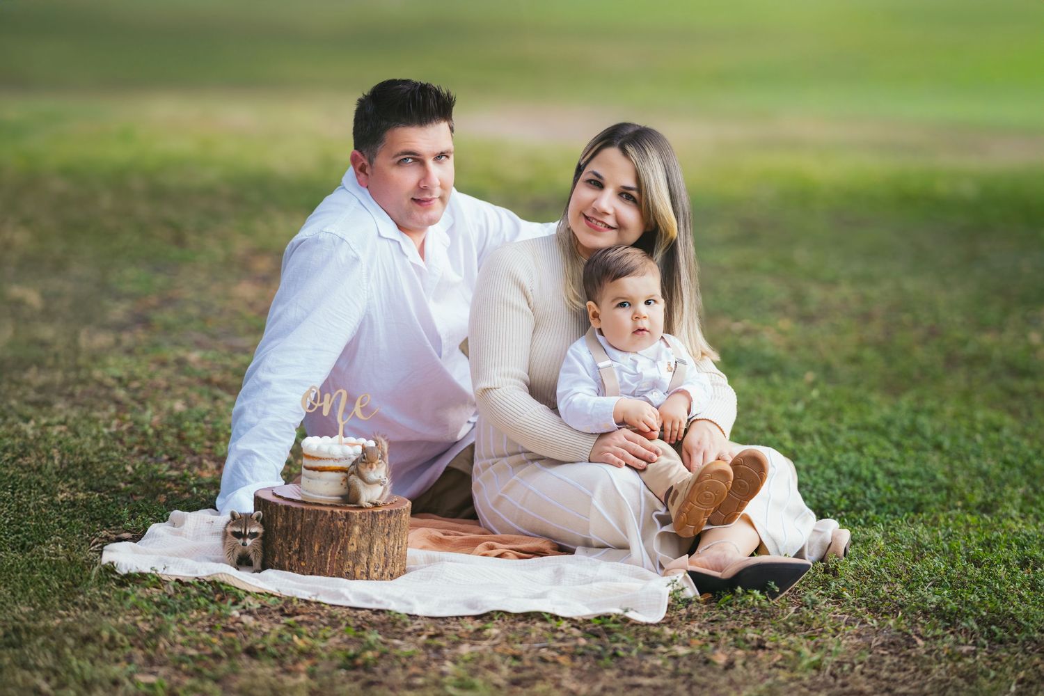 Family of three sitting on a blanket in a park, parents smiling with baby during an outdoor picnic photo session.