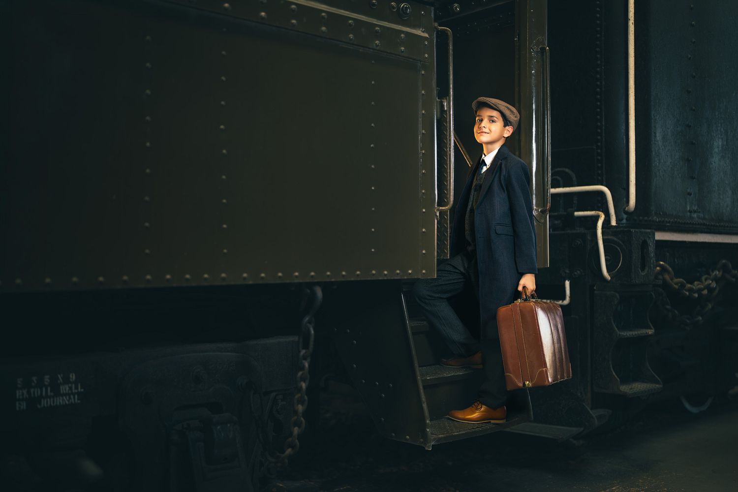A young boy in a blue suit and hat holds a vintage suitcase while standing on train steps in a dark station.