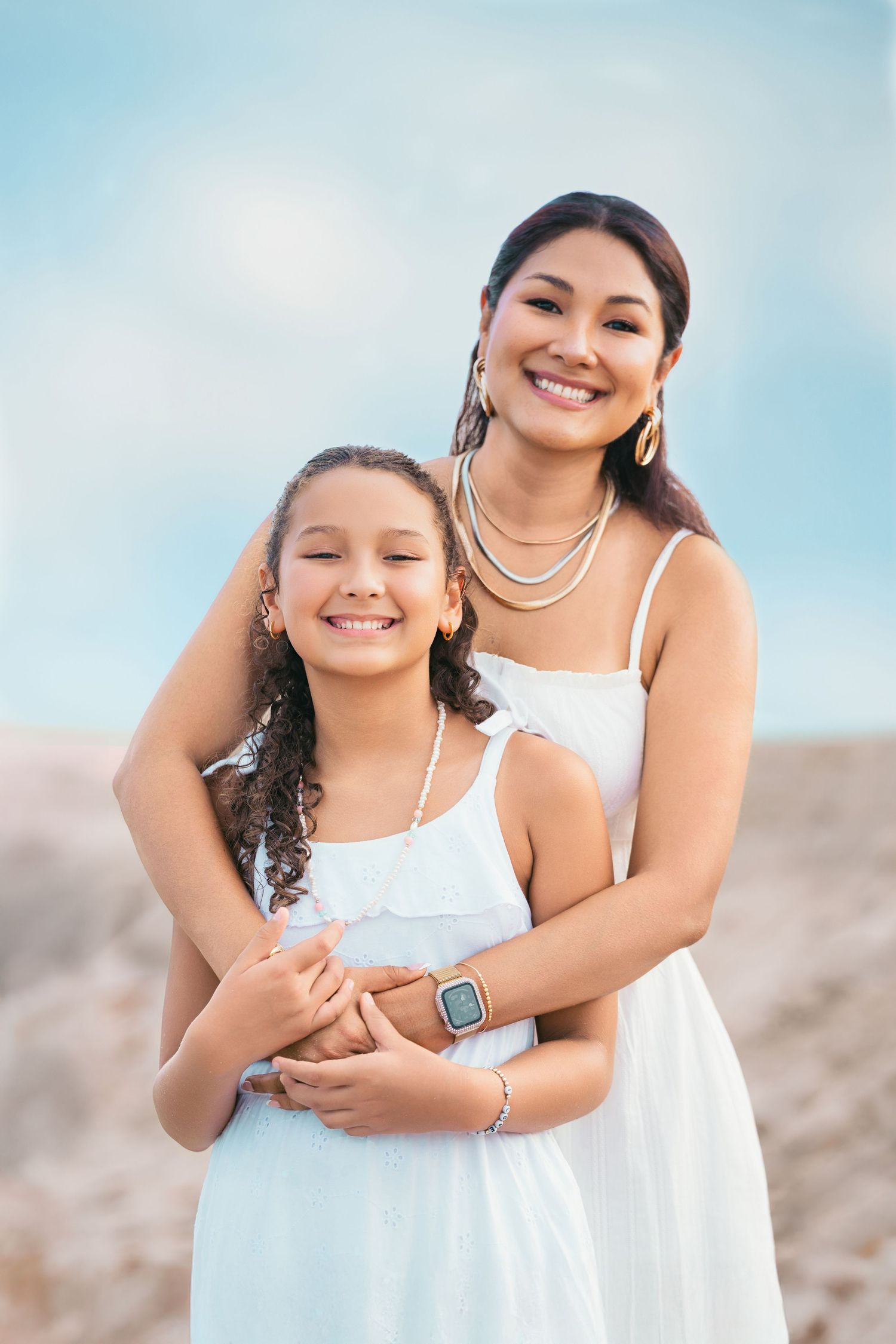 A smiling woman in a white dress hugs a young girl in a matching white dress outdoors against a blue sky.