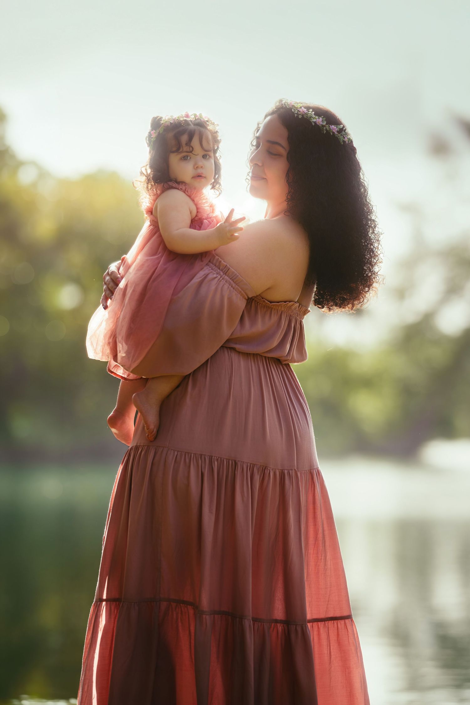 Mother in pink dress holds toddler girl outdoors near water with soft sunlight glowing behind them.