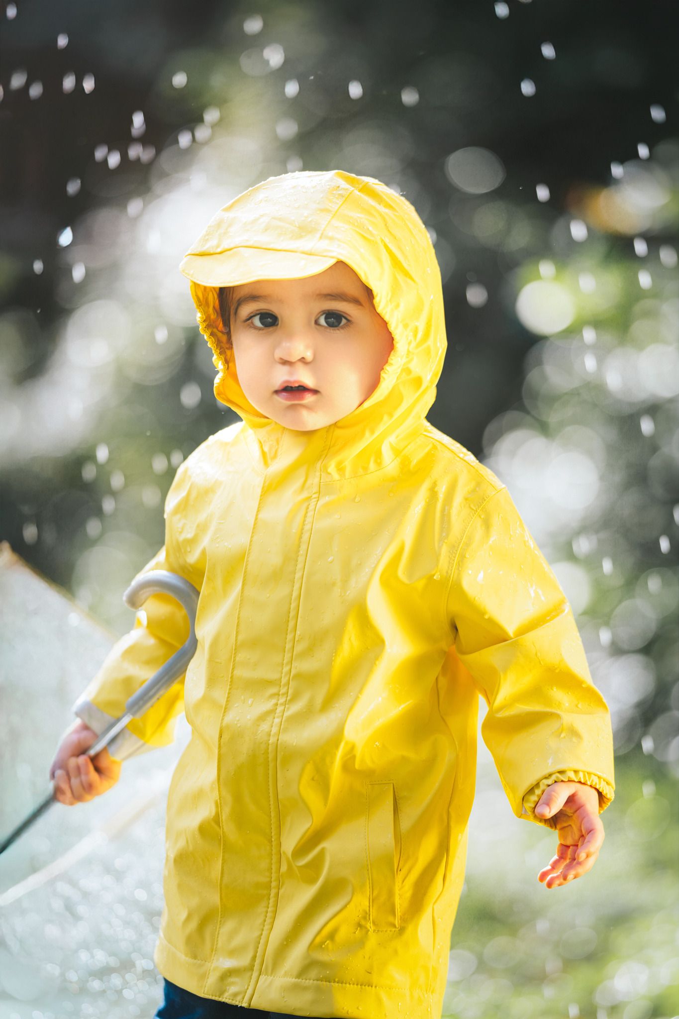 Toddler in yellow raincoat with hood holding umbrella standing in rain outdoors.