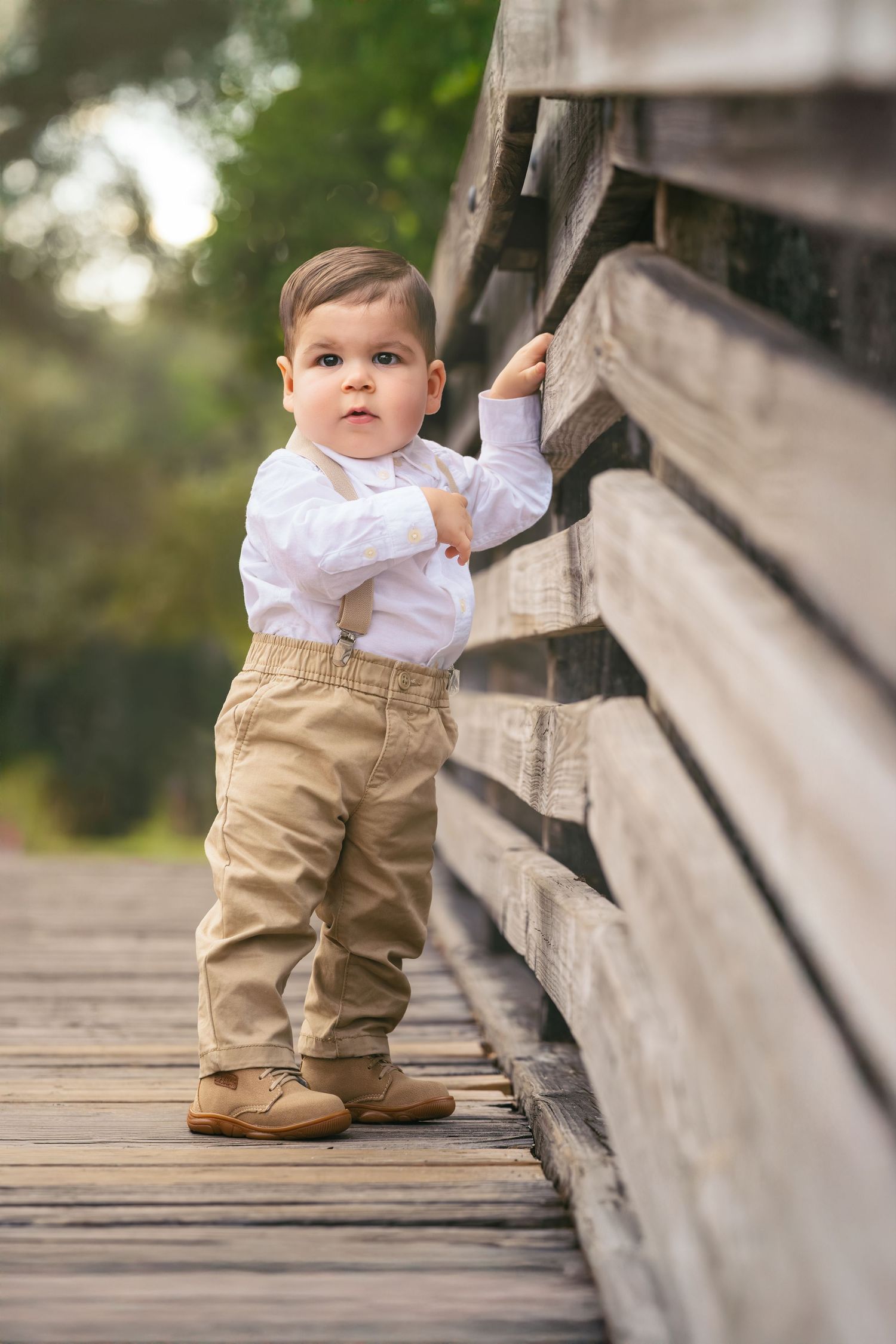 Adorable toddler boy in white shirt and khaki pants standing on a wooden bridge outdoors.