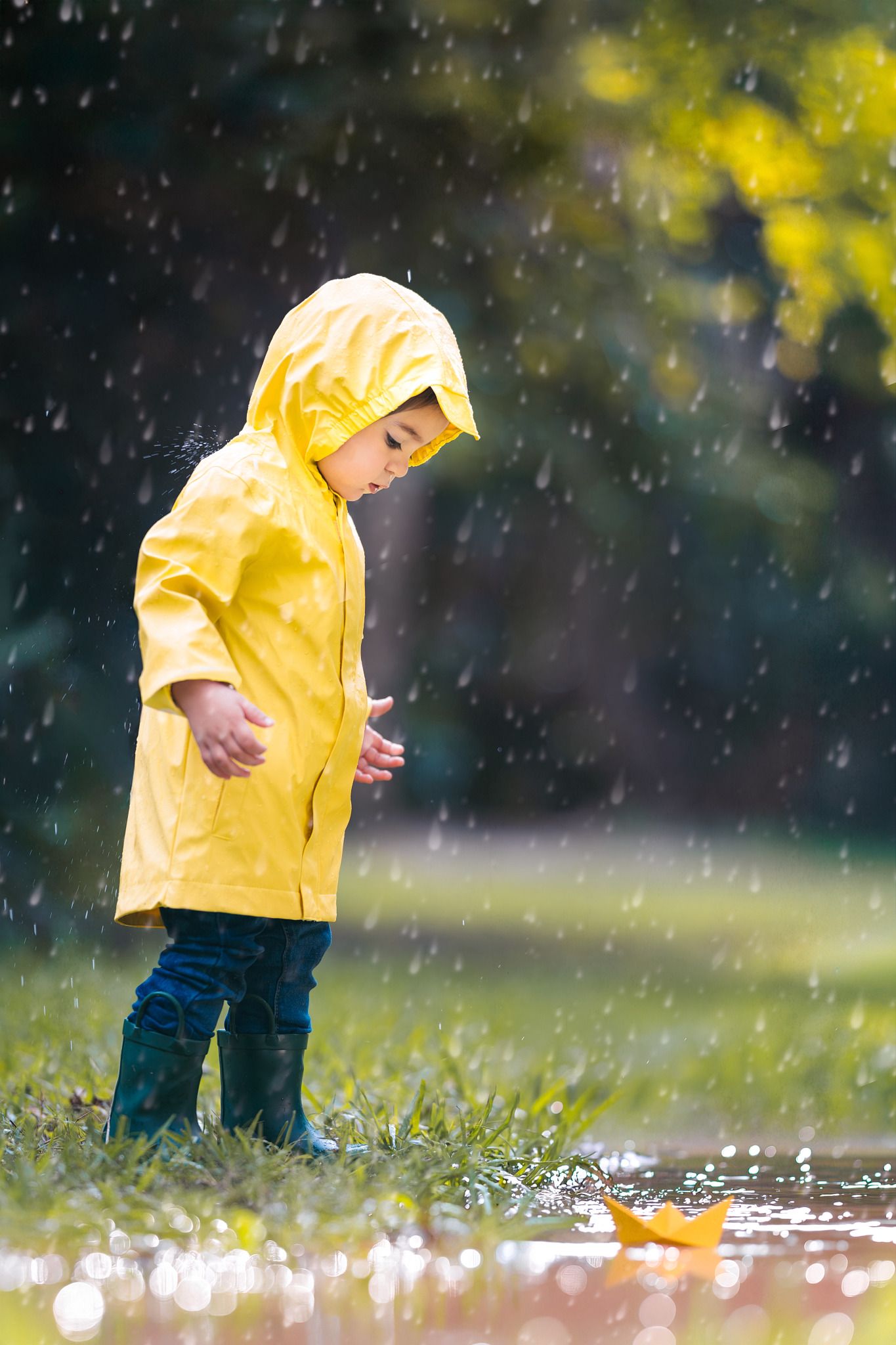 A small child in a yellow raincoat splashes through puddles on a rainy day in the park.