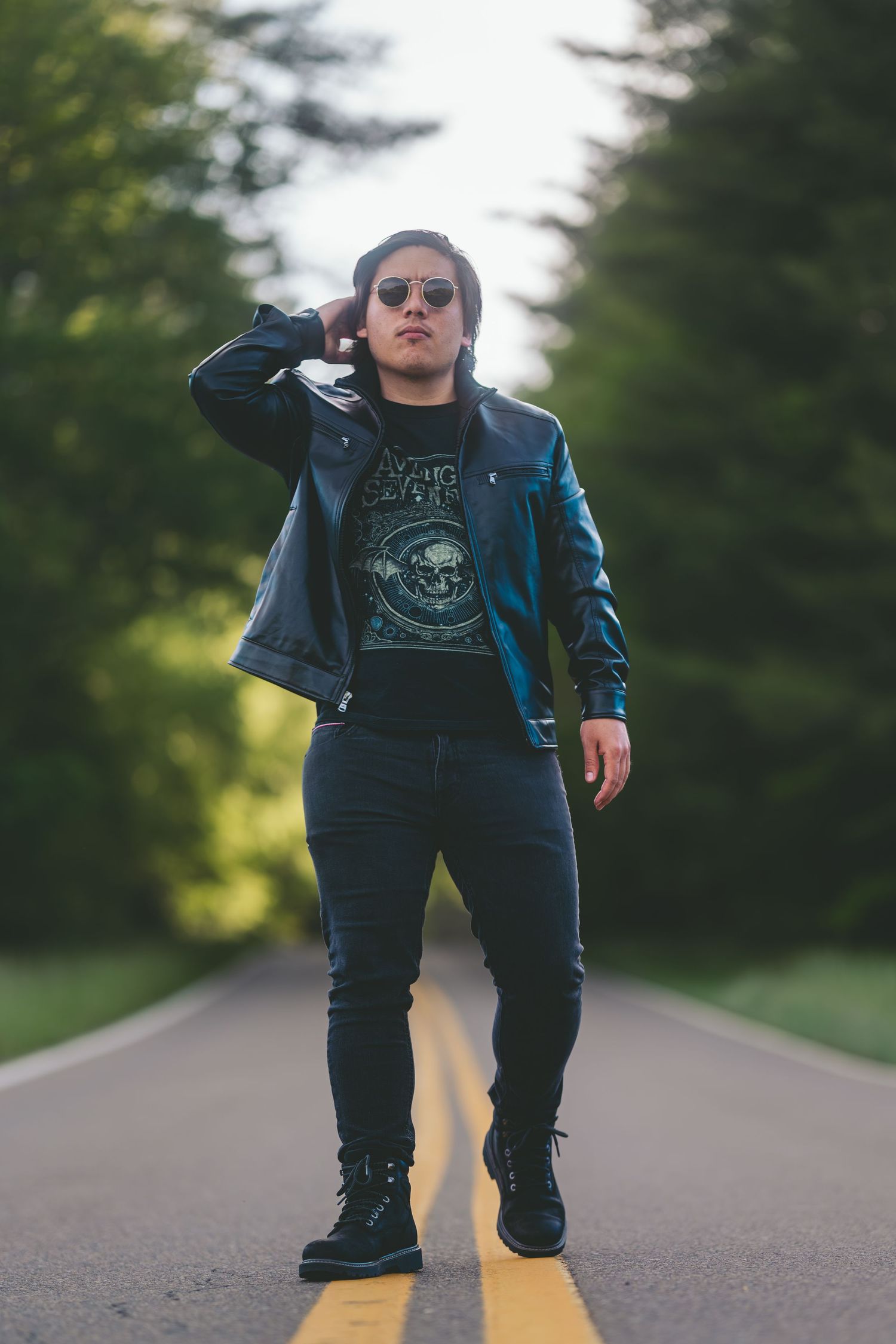A person in dark clothing and sunglasses crouches on a rural road with a blurred forest background.