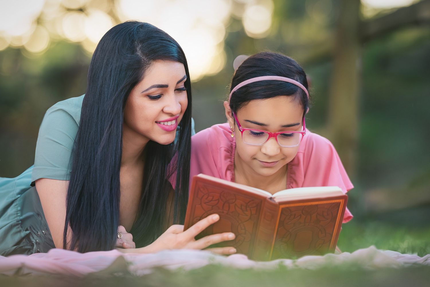 Woman and young girl with glasses reading a book together outdoors in a park setting.