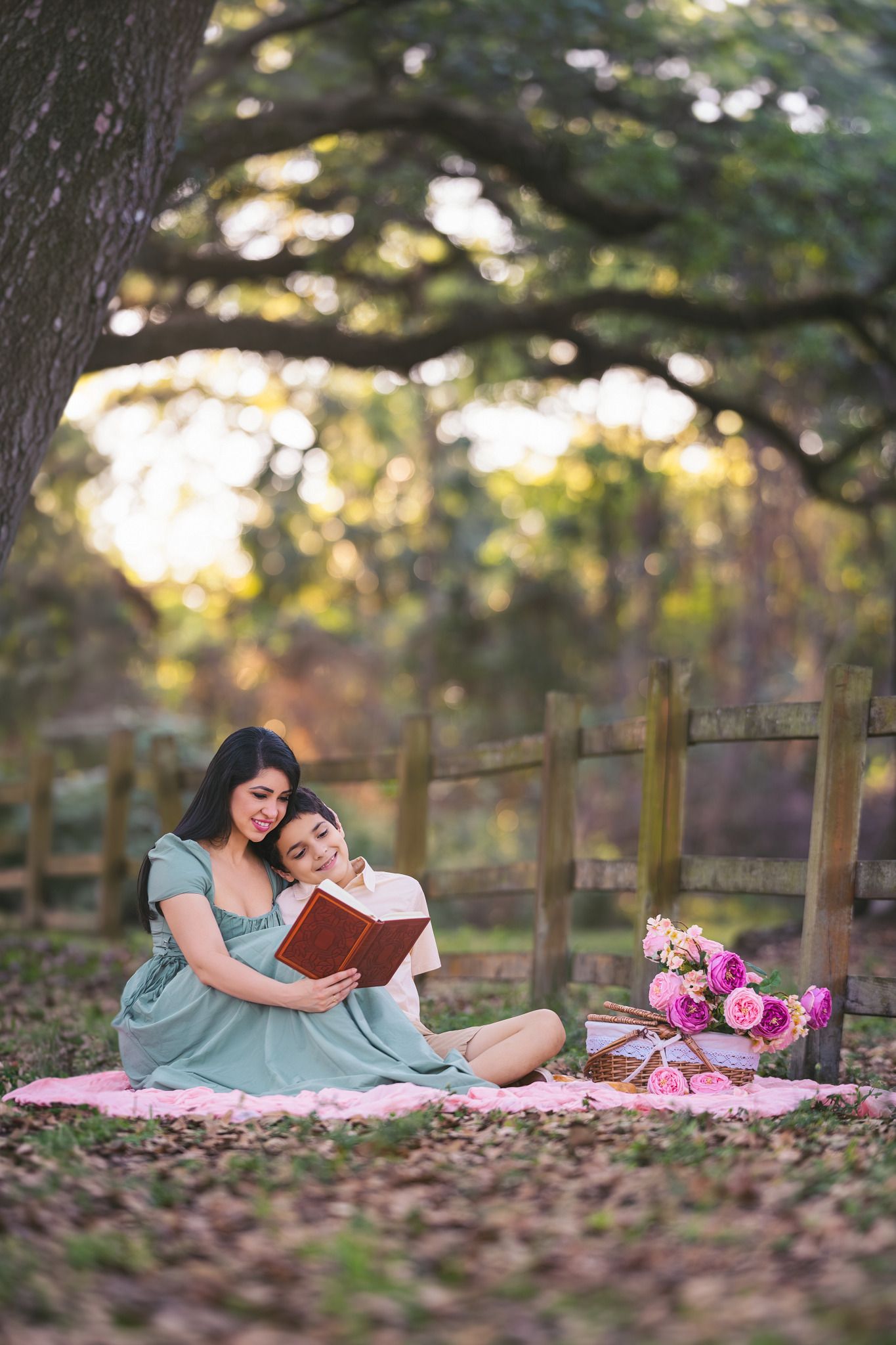 A mother and daughter sit on a pink blanket outdoors, reading a book together near a wooden fence with pink flowers.