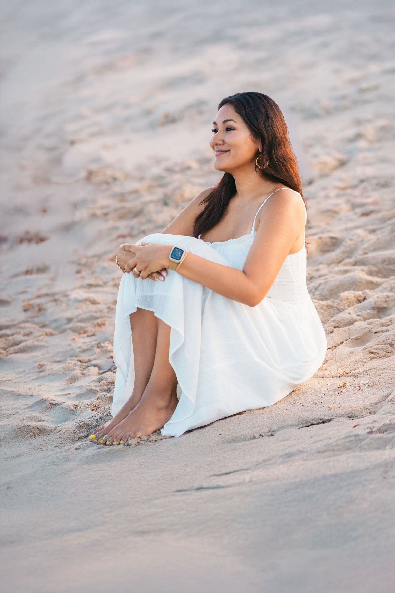 A woman in a flowing white dress reclines on a sandy beach during golden hour.