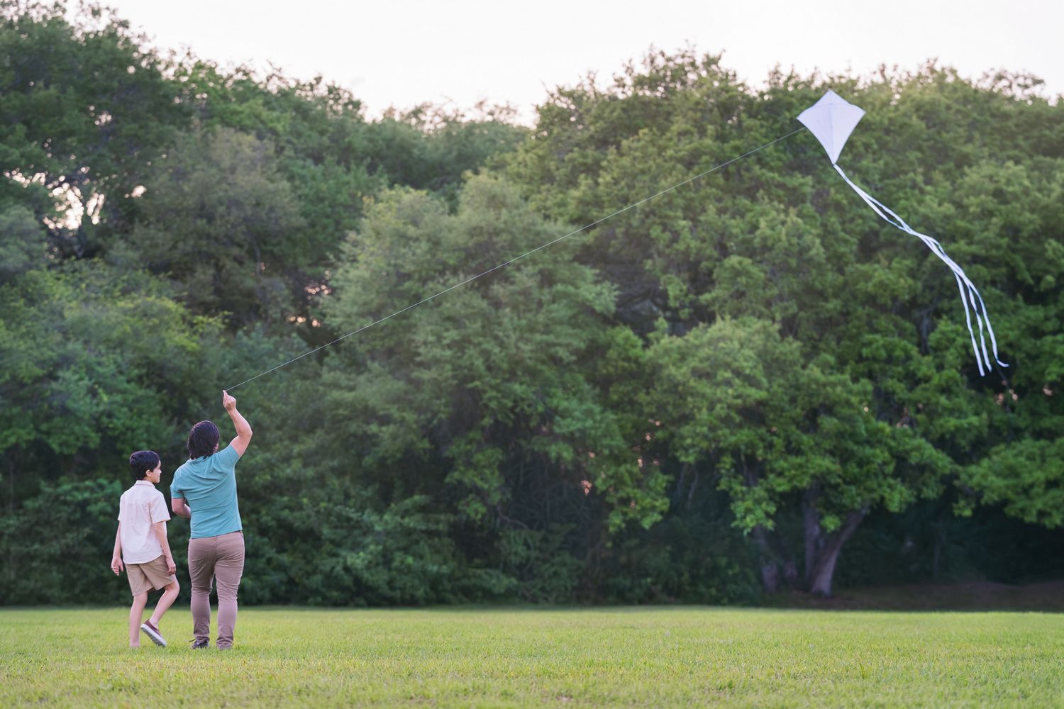 Two people flying a white kite in a green park surrounded by tall trees on a calm evening.