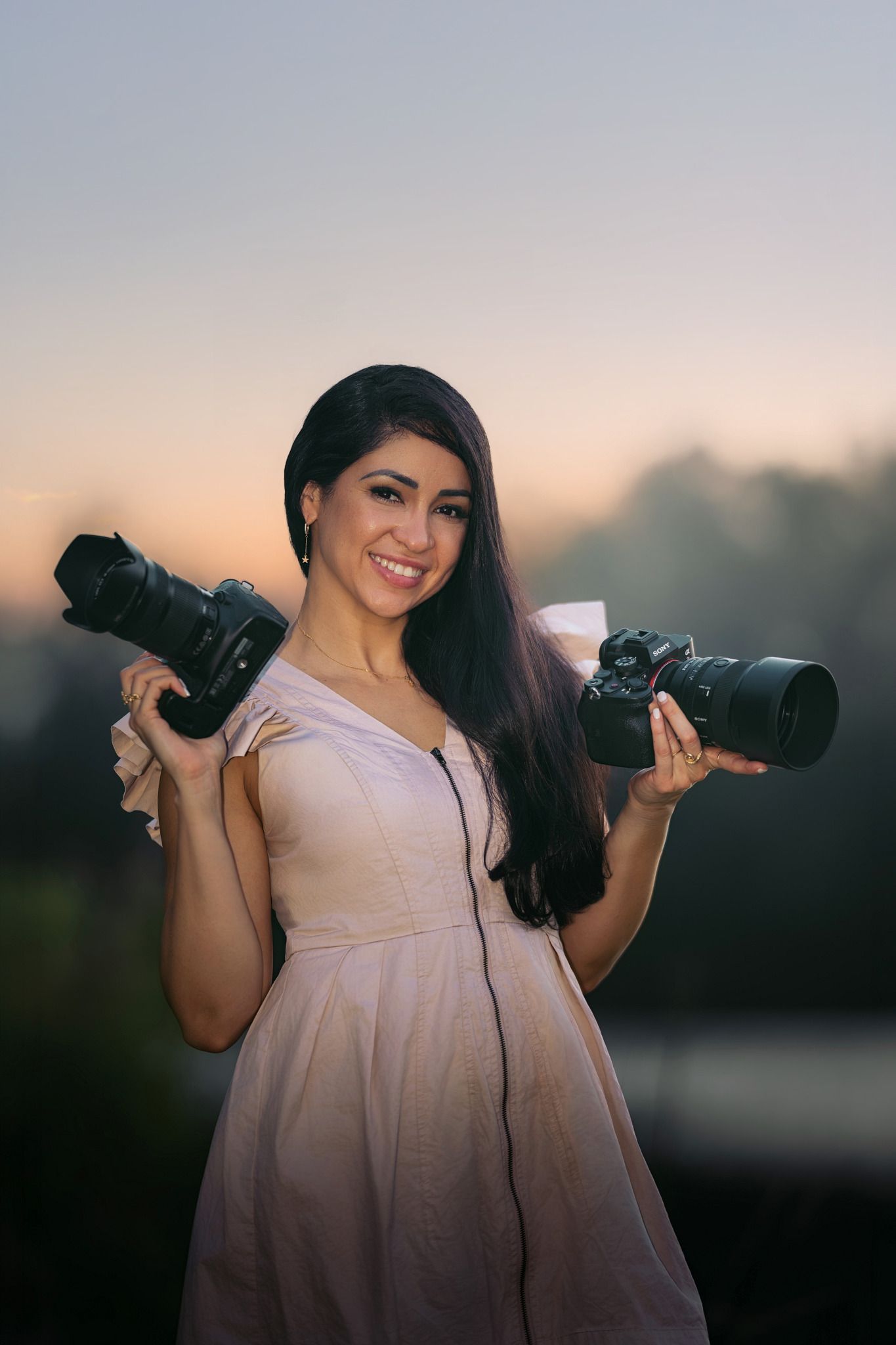 Smiling woman in pink dress holding two professional DSLR cameras outdoors at sunset.