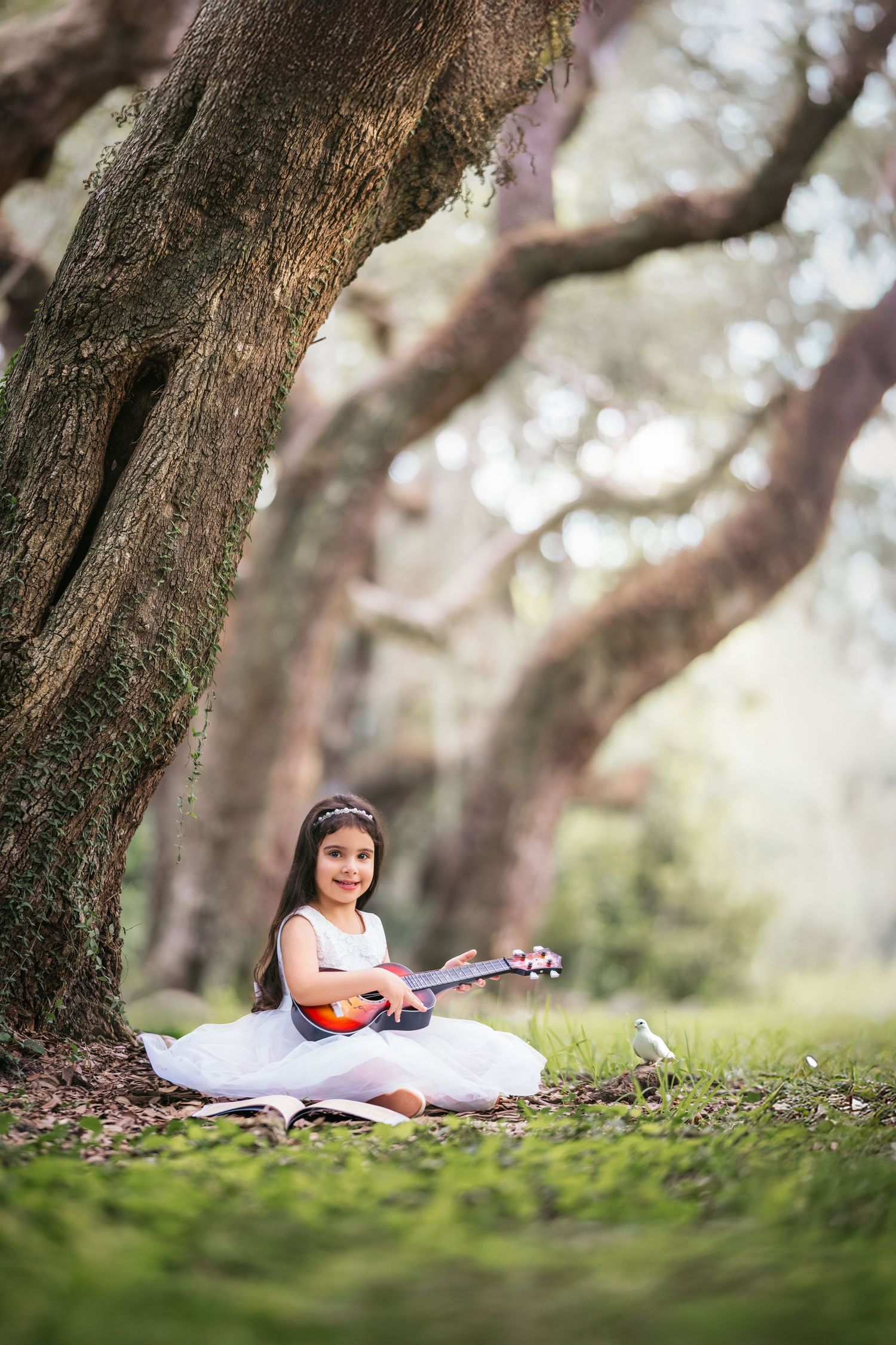 Young girl in white dress playing ukulele under large oak trees in a lush green park setting.