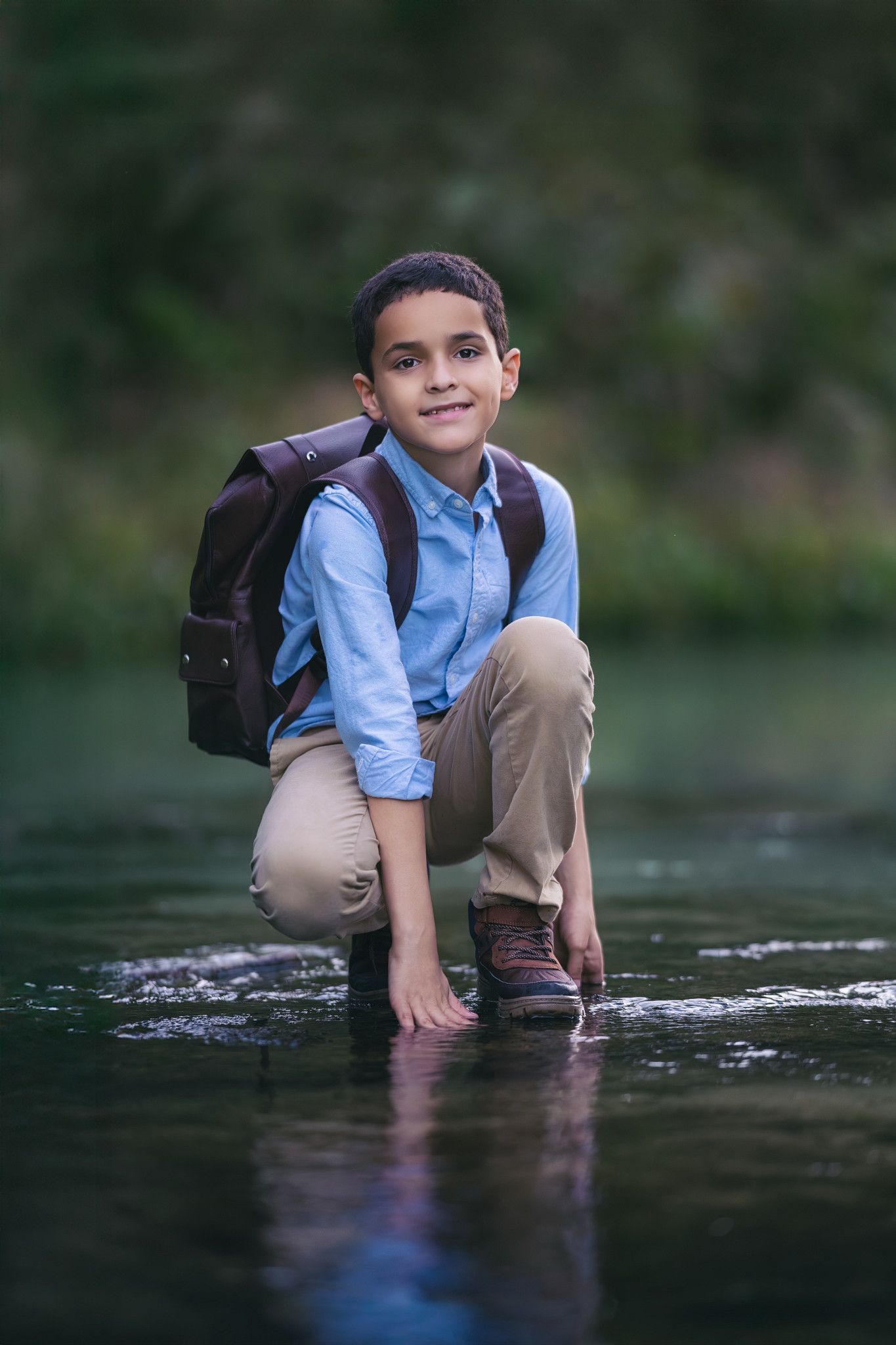 Smiling boy with backpack crouching in a shallow river, wearing a blue shirt and khaki pants outdoors.