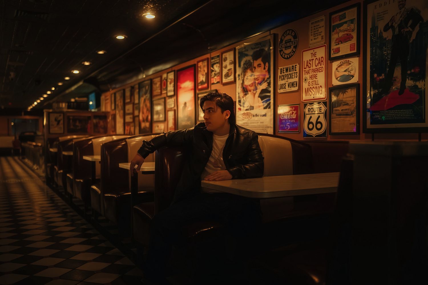 Young man in black leather jacket sitting alone in a vintage diner with Route 66 signs and movie posters.
