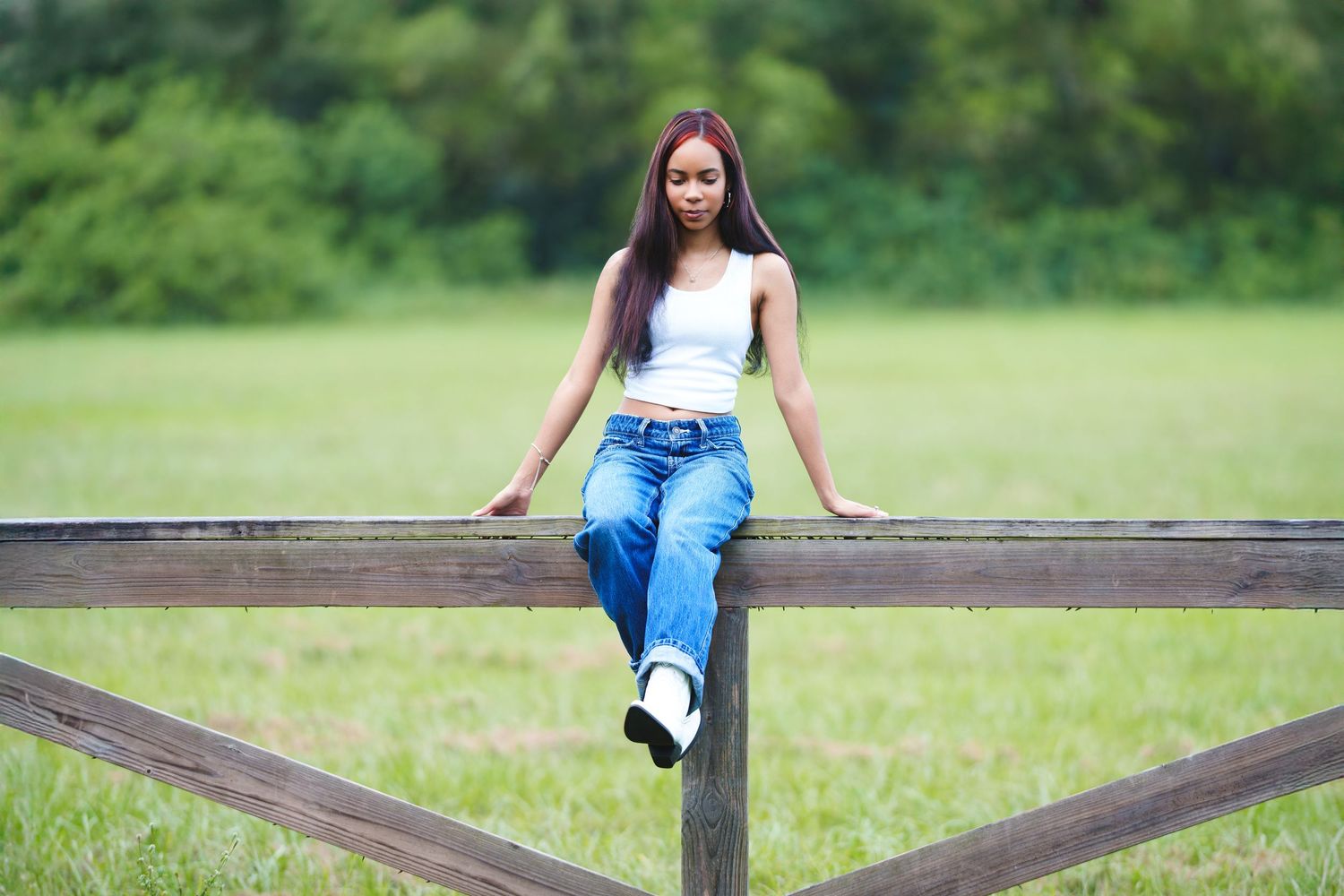 Woman sitting on a wooden fence in a green outdoor setting.