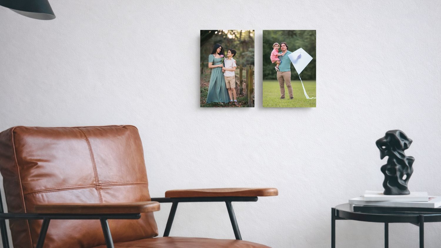 Two canvas prints of family photos displayed on a white wall above a leather armchair and side table with a sculpture.