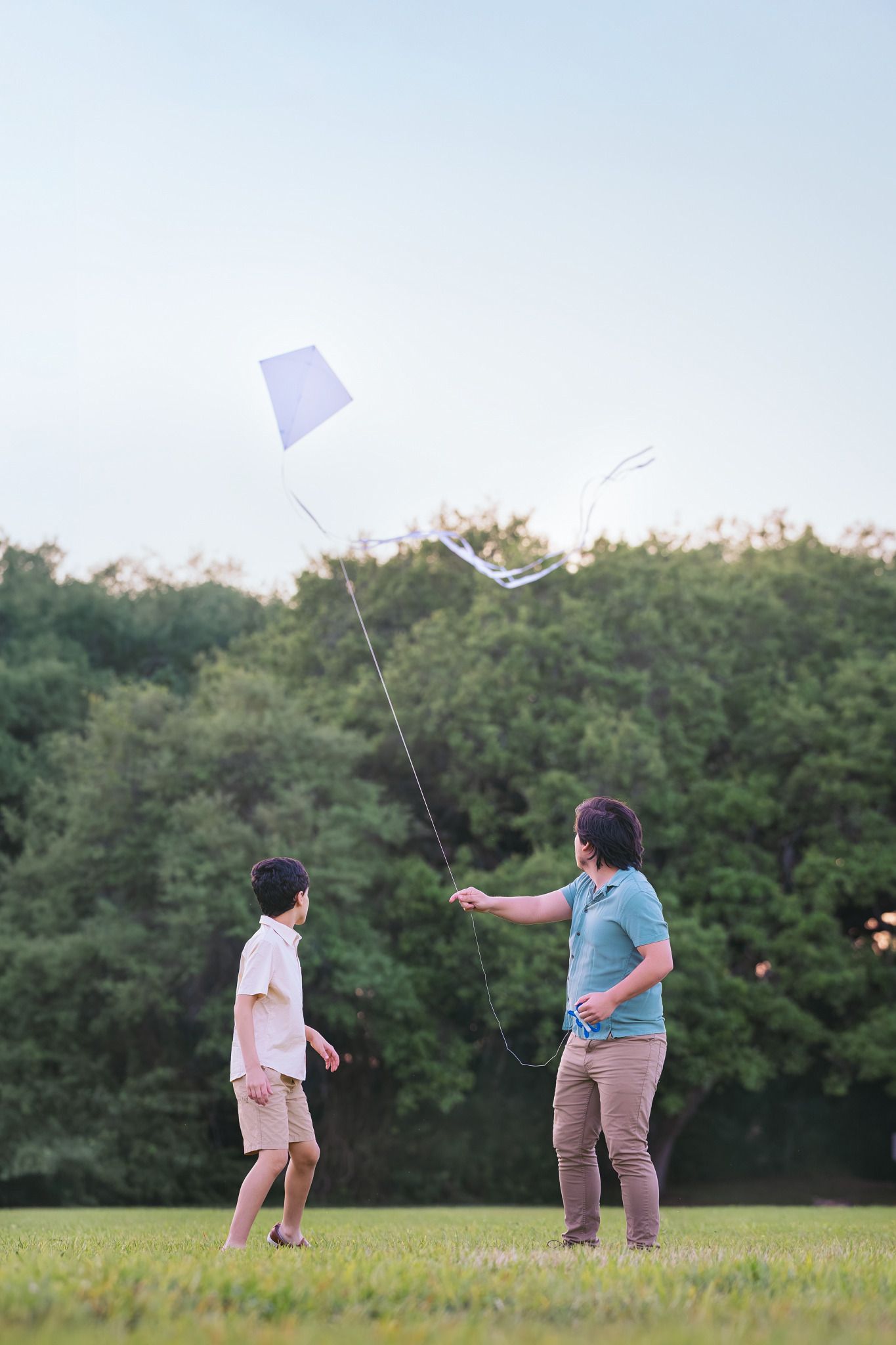 Father and son flying a white kite together in a green park on a clear evening.
