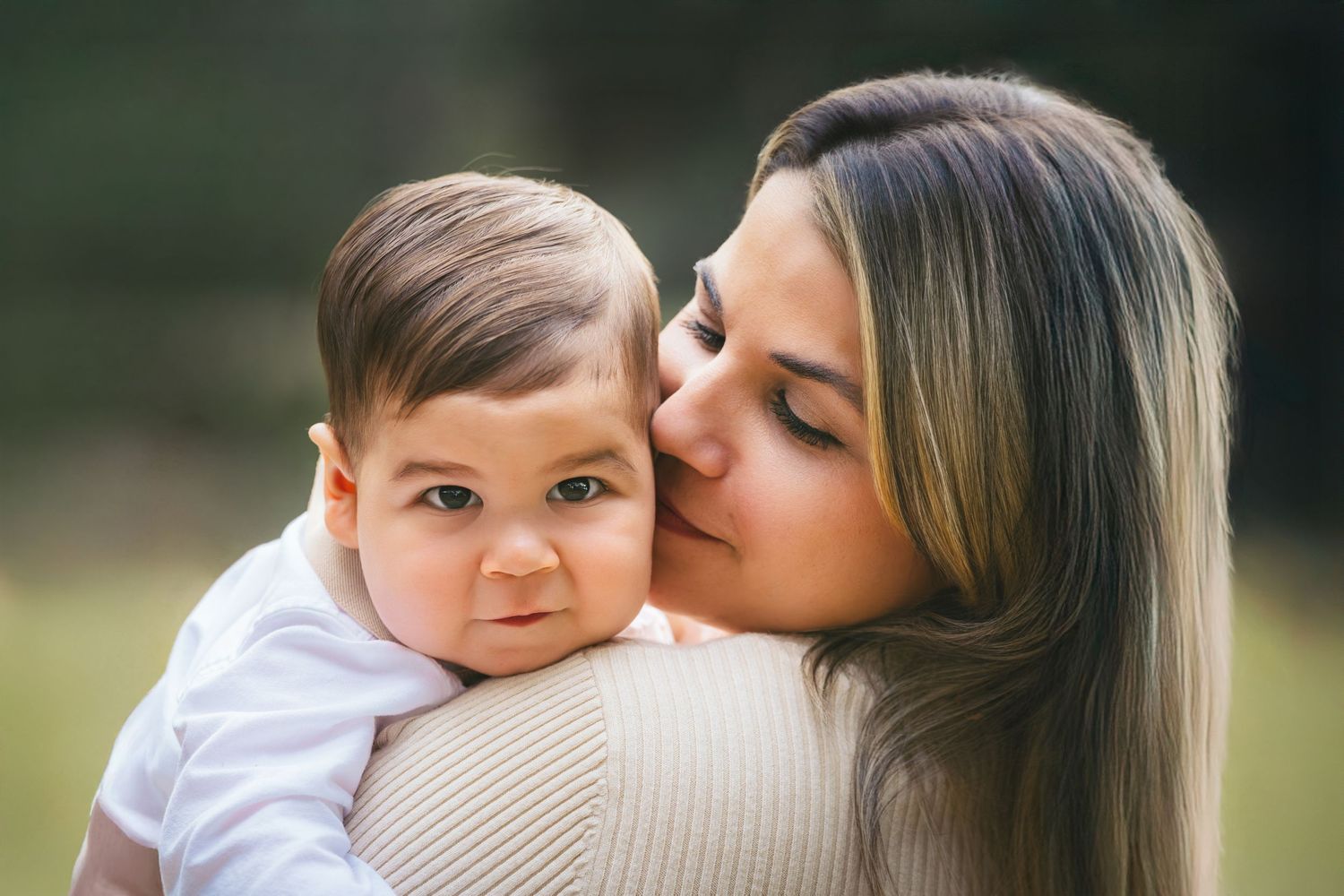 A loving mother kissing her baby boy on the cheek while holding him outdoors in soft natural light.