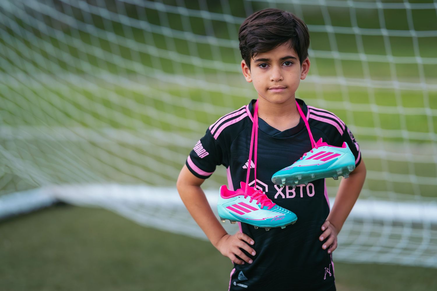 Young soccer player in black XBTO jersey holding pink and blue Adidas cleats in front of a goal net.