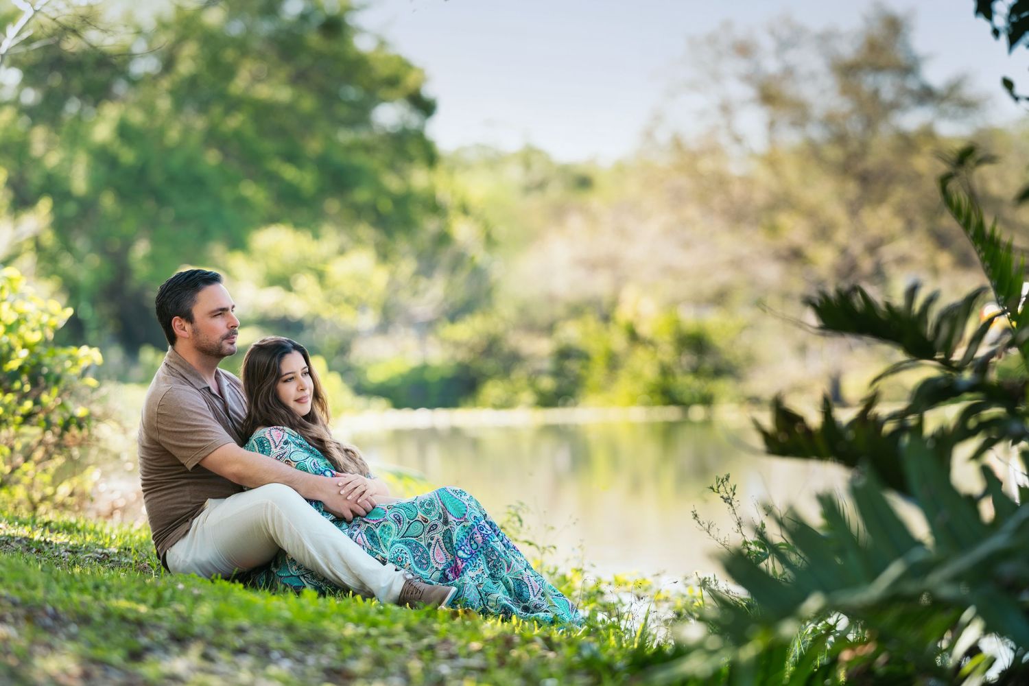 Couple sitting together on grass by a serene lake surrounded by lush green trees in a peaceful park setting.