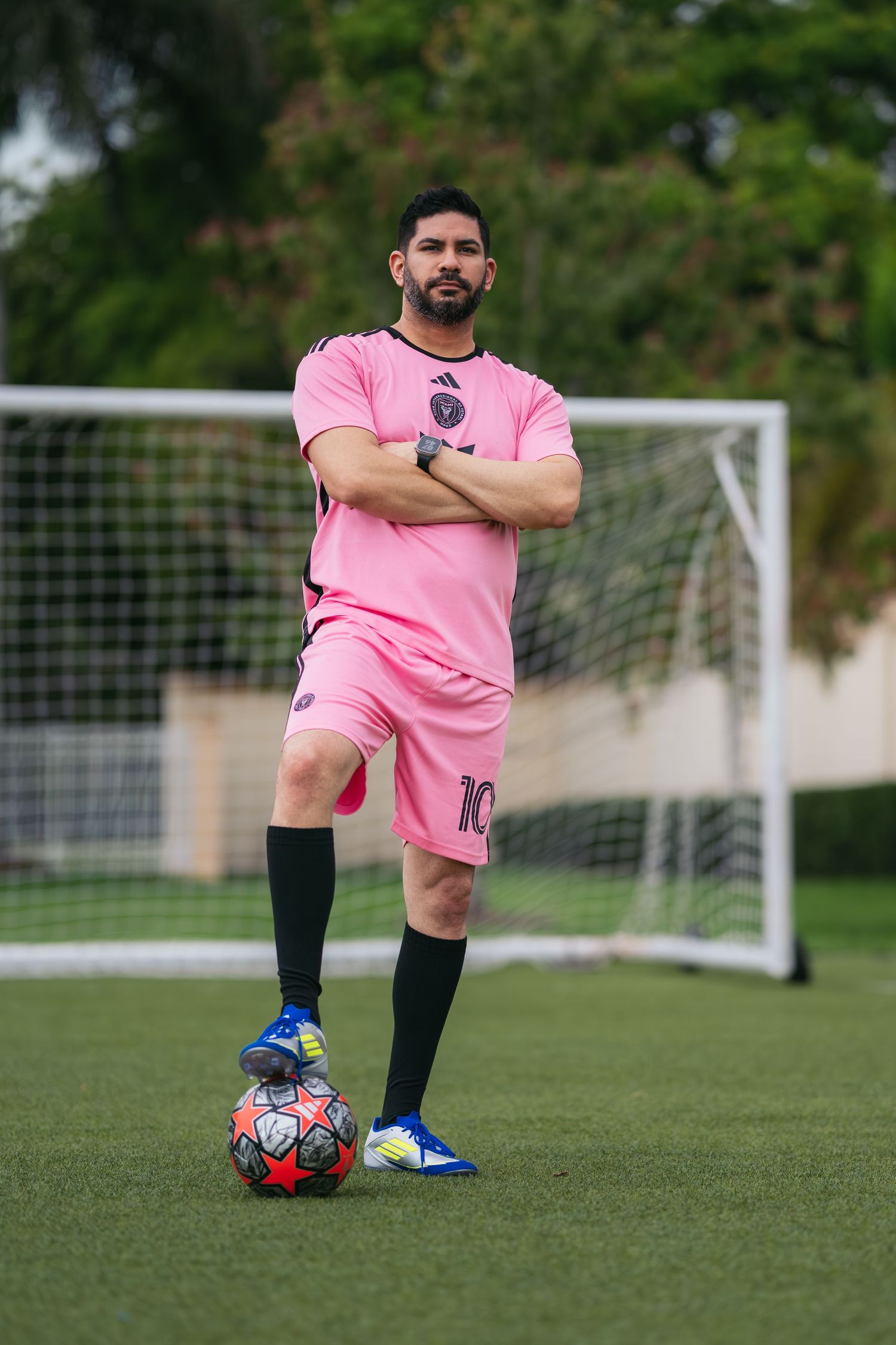 Man in pink soccer jersey number 10 stands confidently with foot on ball near goal post on green field.