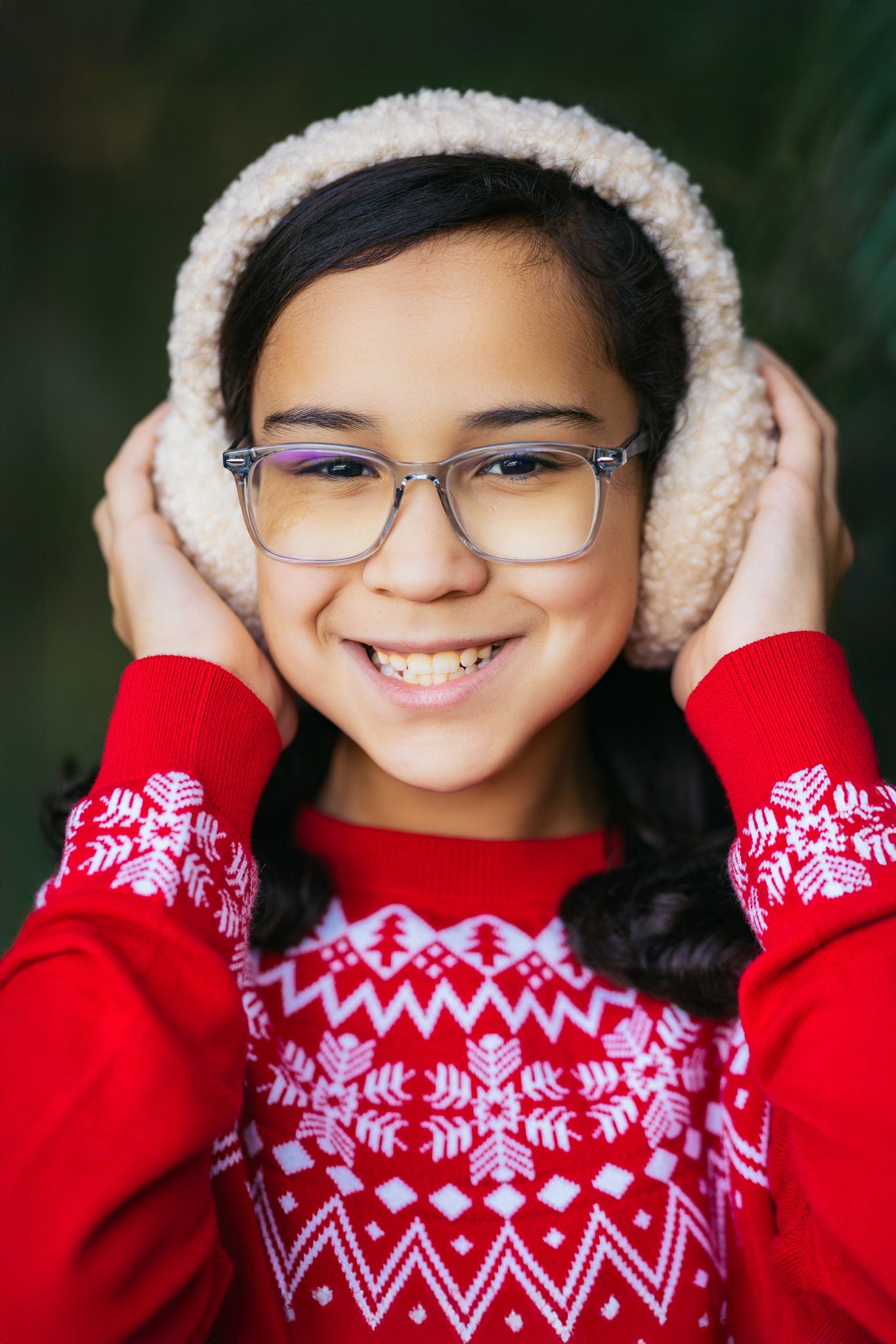 Smiling girl in red Nordic sweater and fuzzy earmuffs wearing glasses, posing outdoors in winter.