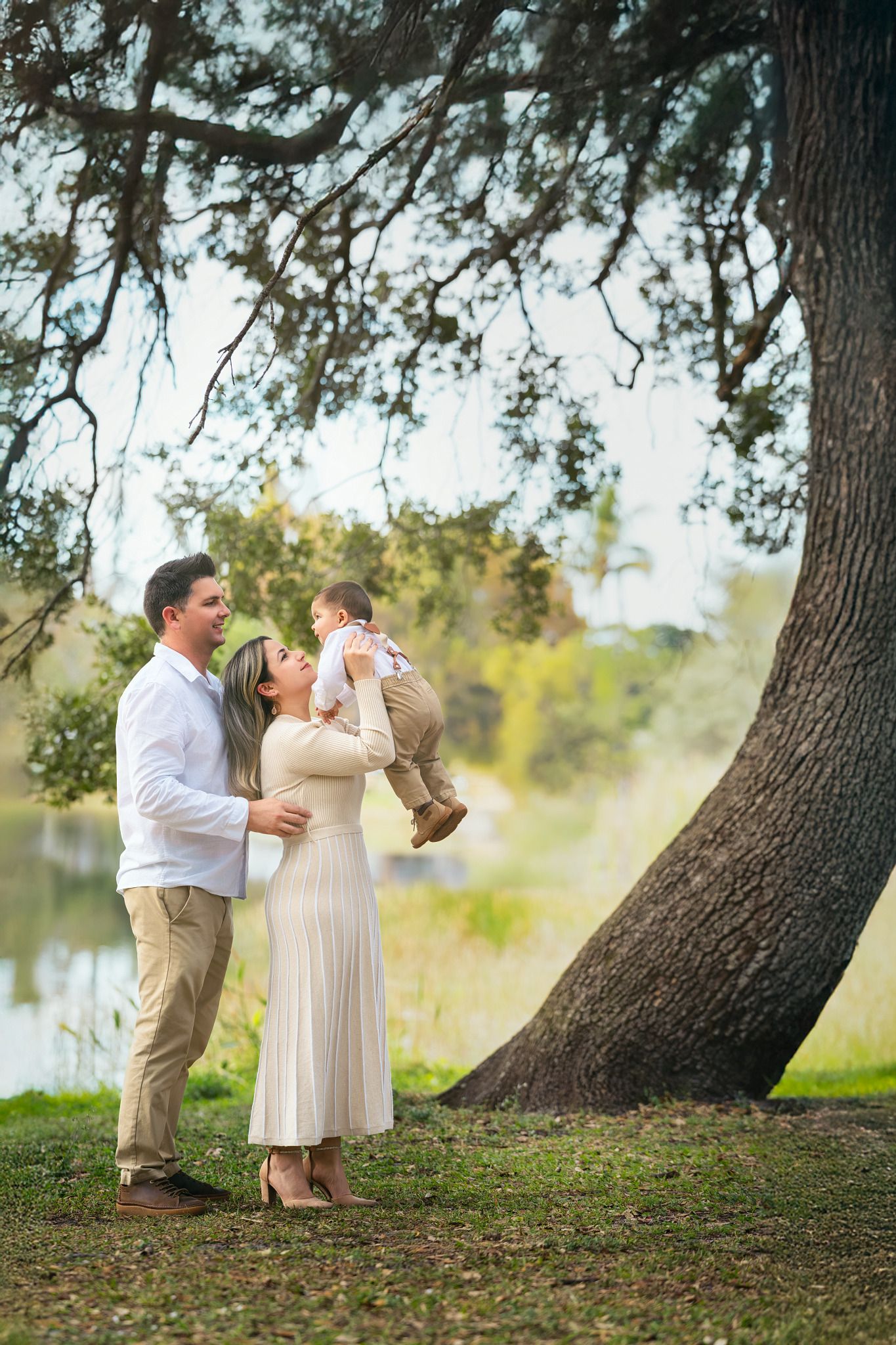 A romantic couple in light-colored attire shares tender moments under large pine trees by a lake.