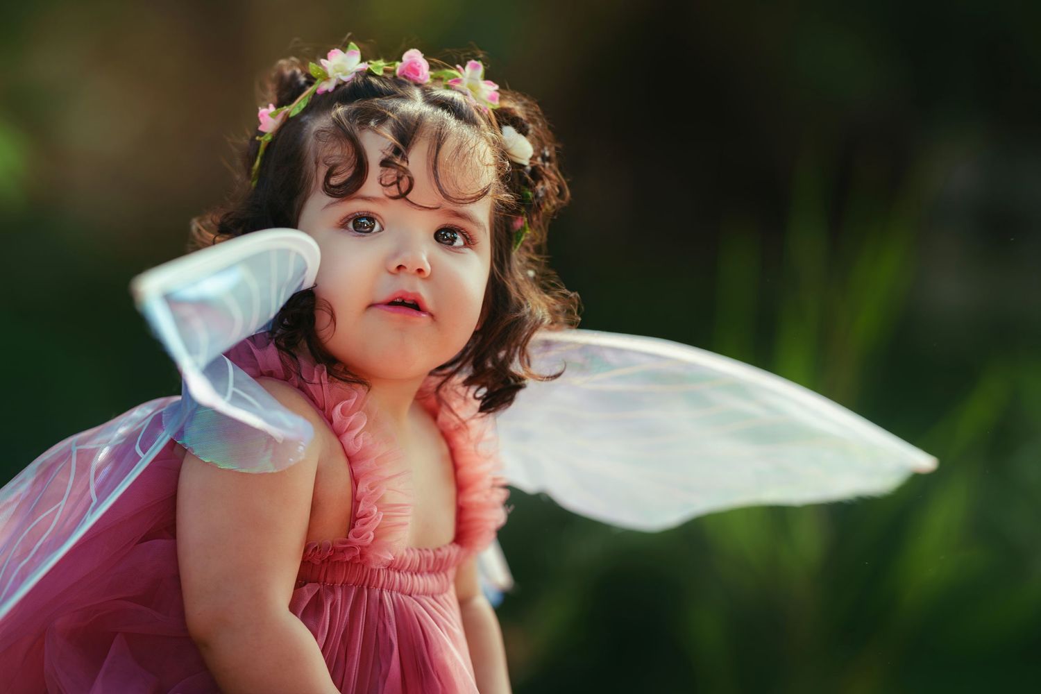 Adorable toddler girl dressed as a fairy in pink dress with wings and floral crown outdoors.