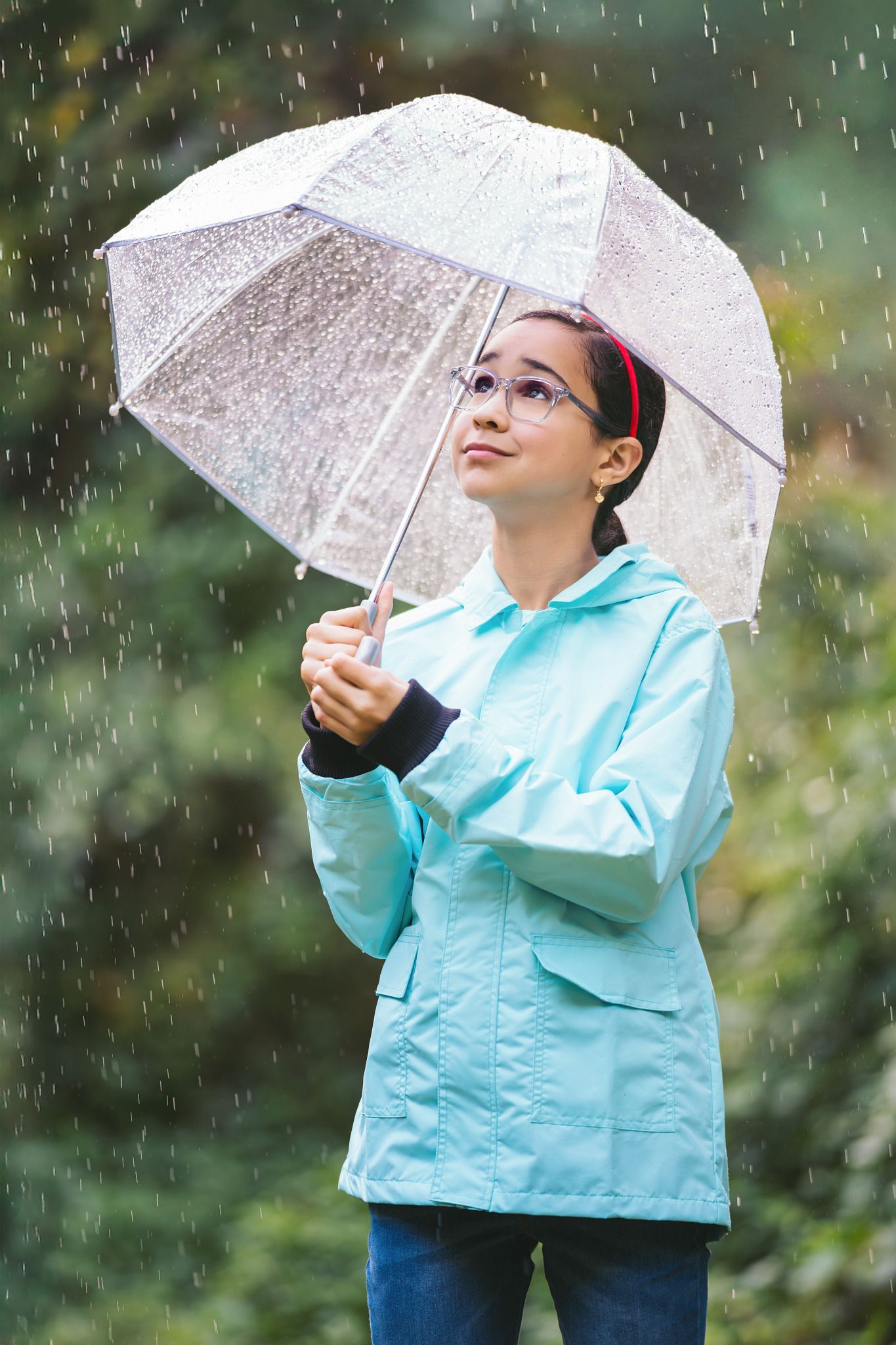 Young girl in teal raincoat holding clear umbrella in rain, looking up at sky outdoors.