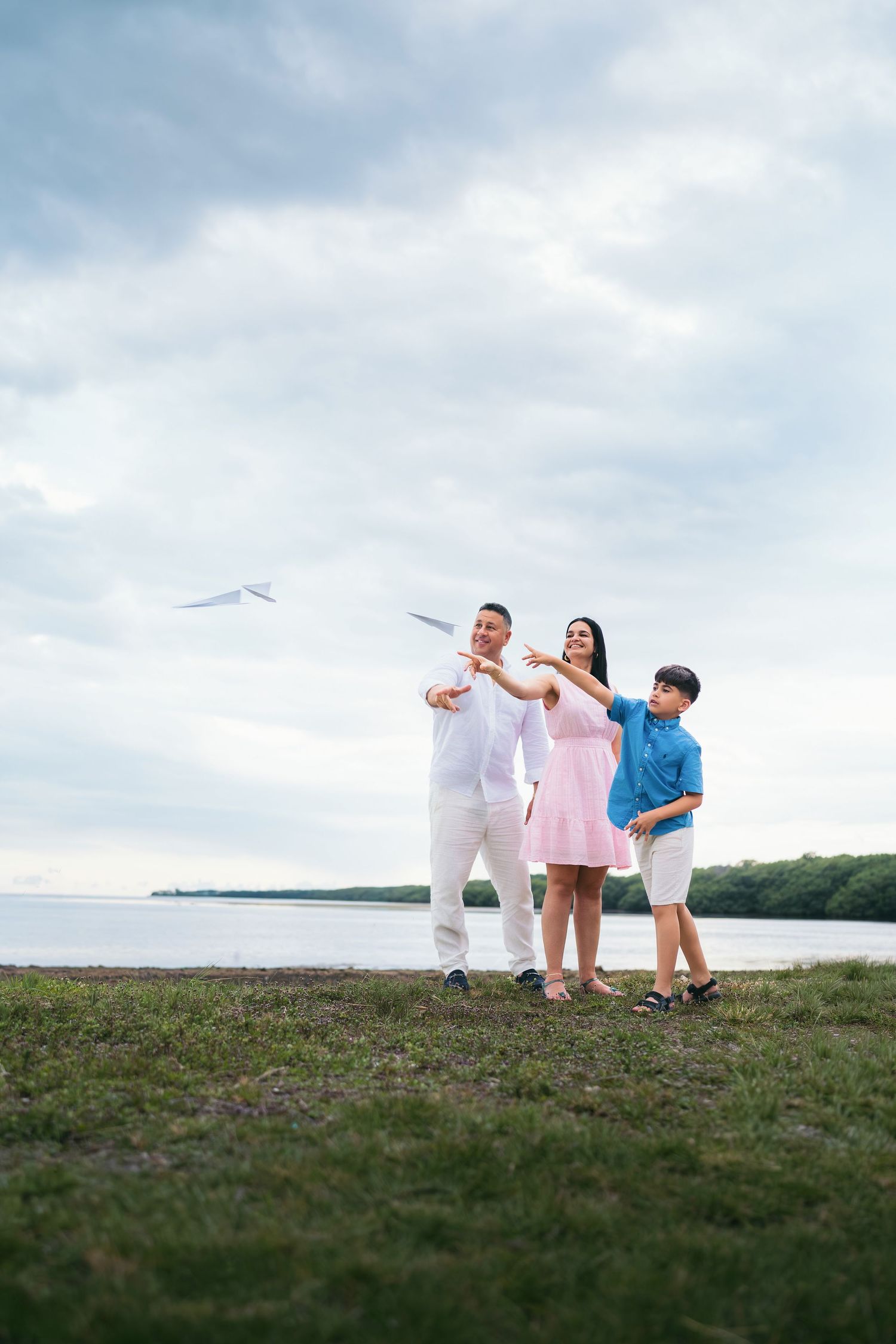 Happy family of three launching a paper airplane by the lake on a cloudy day.