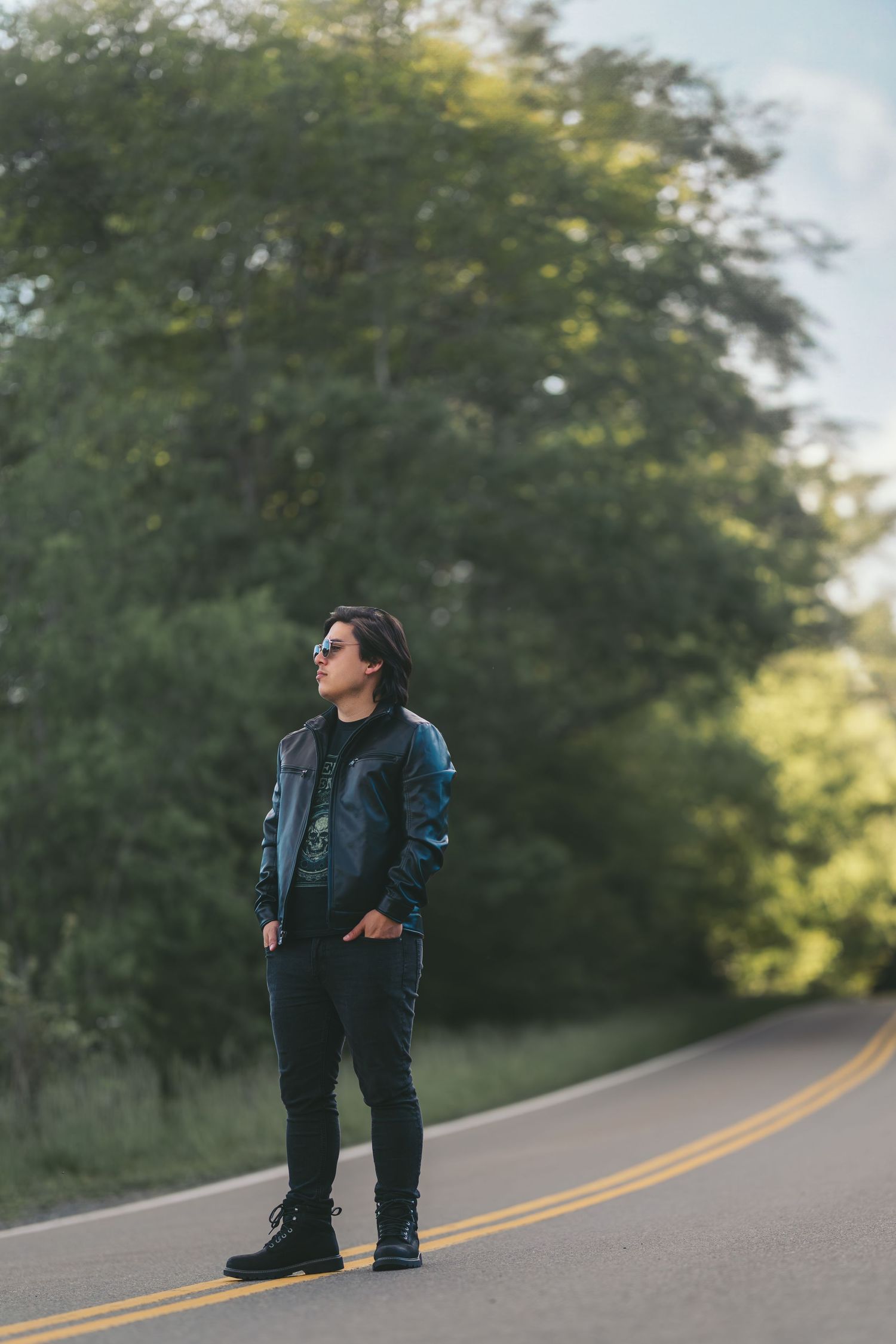 A person in dark clothing stands on a winding road surrounded by lush green trees in natural lighting.