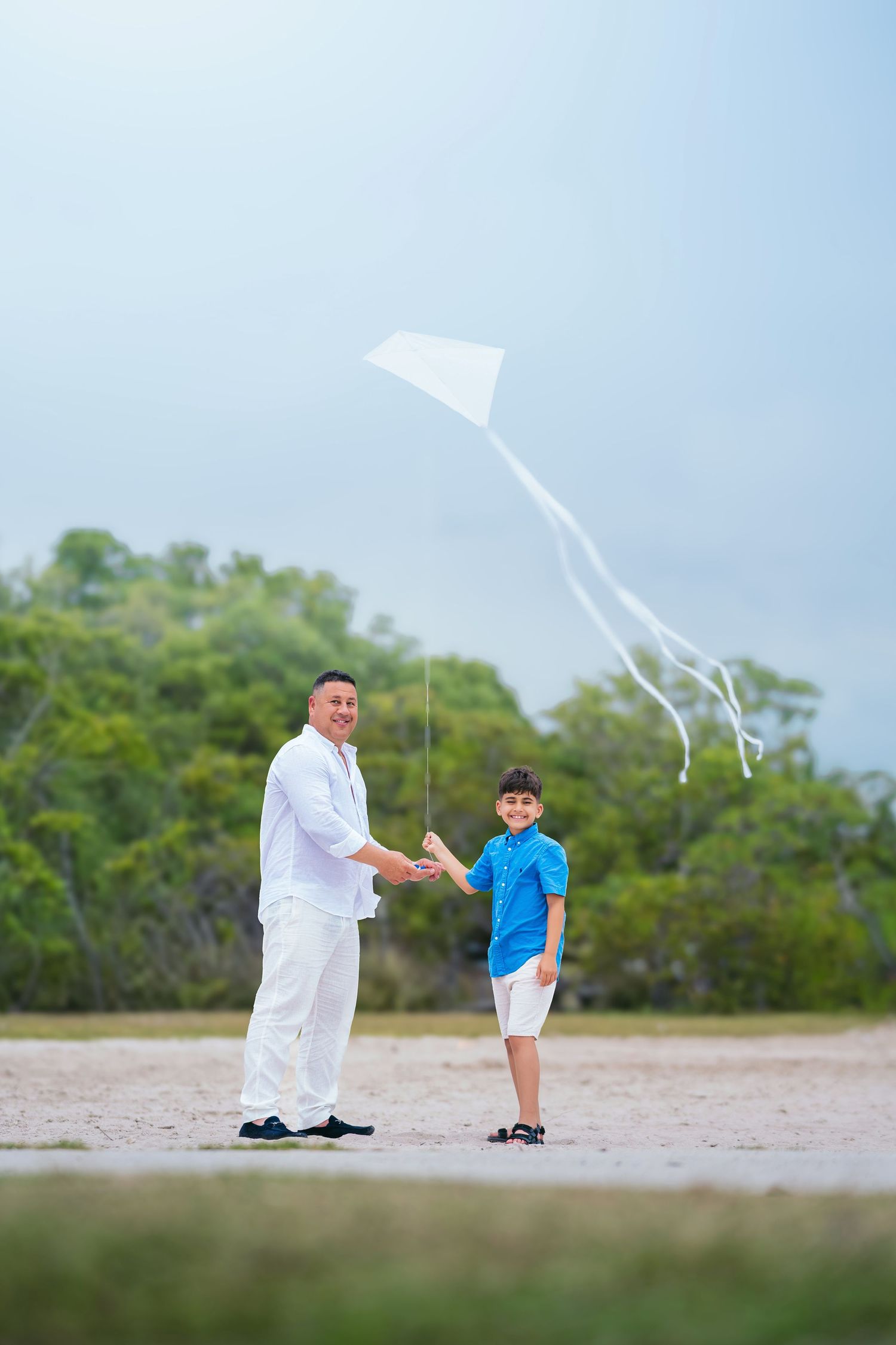 Father and son flying a white kite together on a sandy beach with green trees in the background.