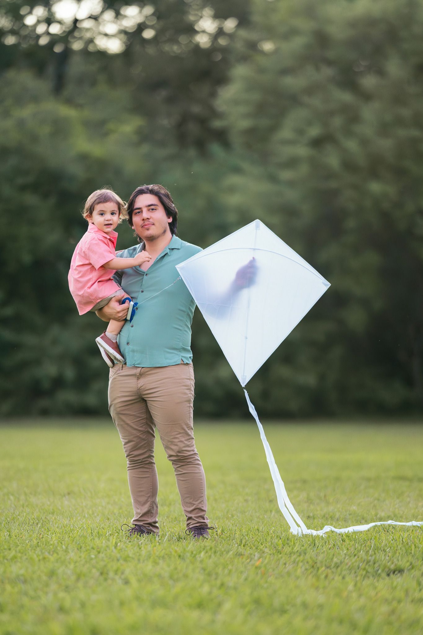 Father holding young child while flying a white kite in a green park on a sunny day.