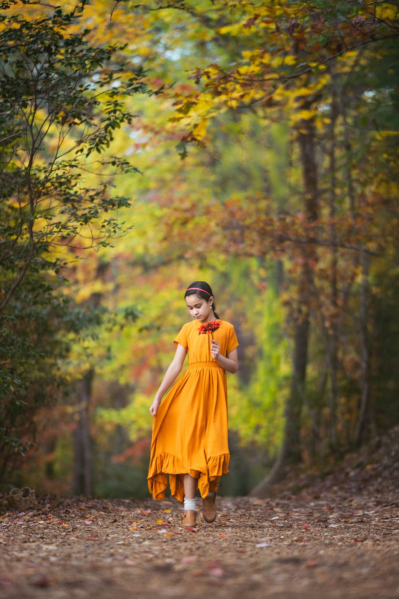 Person in flowing orange dress walking along a forest path covered in autumn leaves.