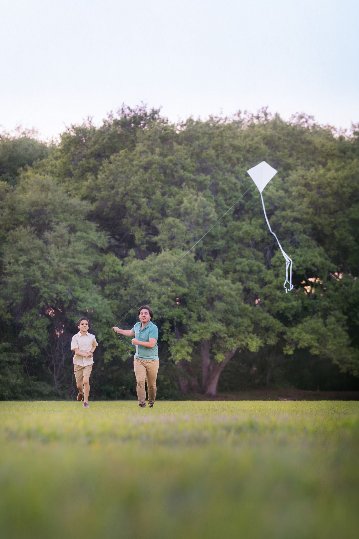 Two people running and flying a white kite in a green park with trees in the background.