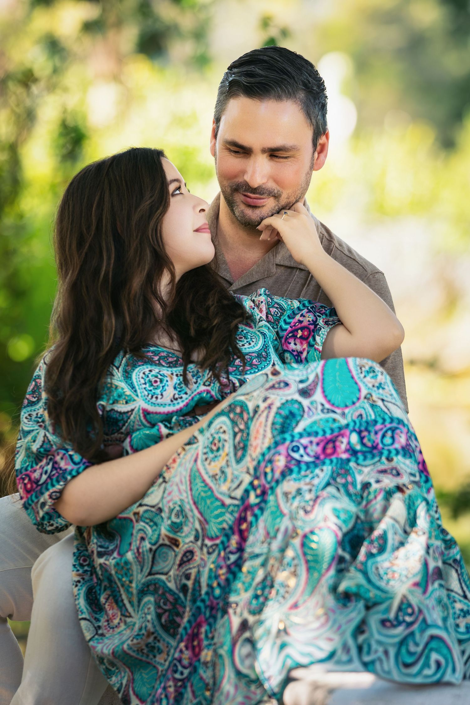 A couple shares a tender moment outdoors, woman in paisley dress gazing up at man holding her in a lush green setting.