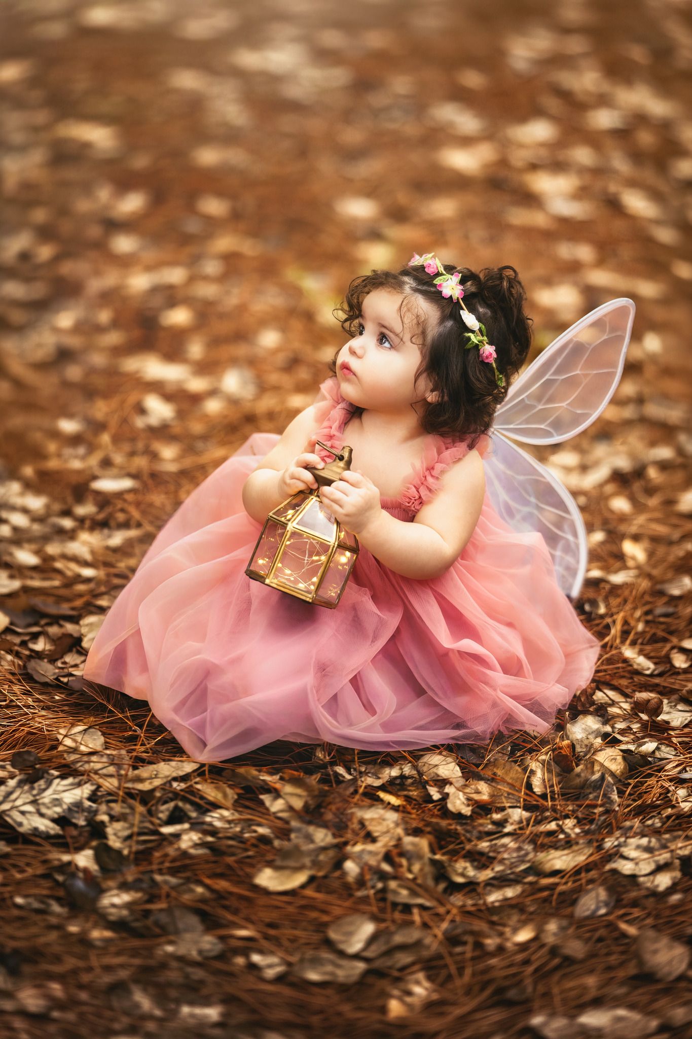 A toddler girl in a pink tutu dress with fairy wings and floral crown holds a golden lantern outdoors.
