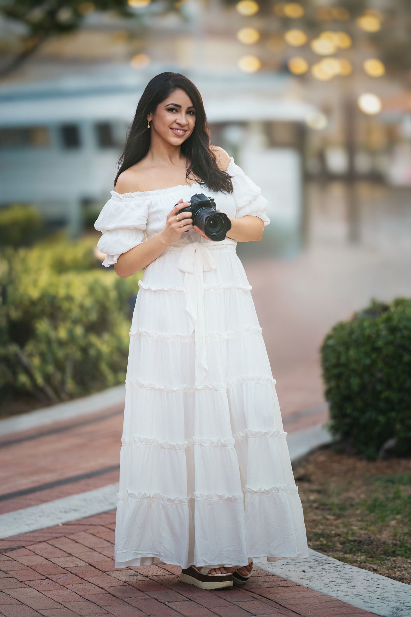 A person in a flowing white off-shoulder dress poses outdoors in a garden setting with soft bokeh lighting.