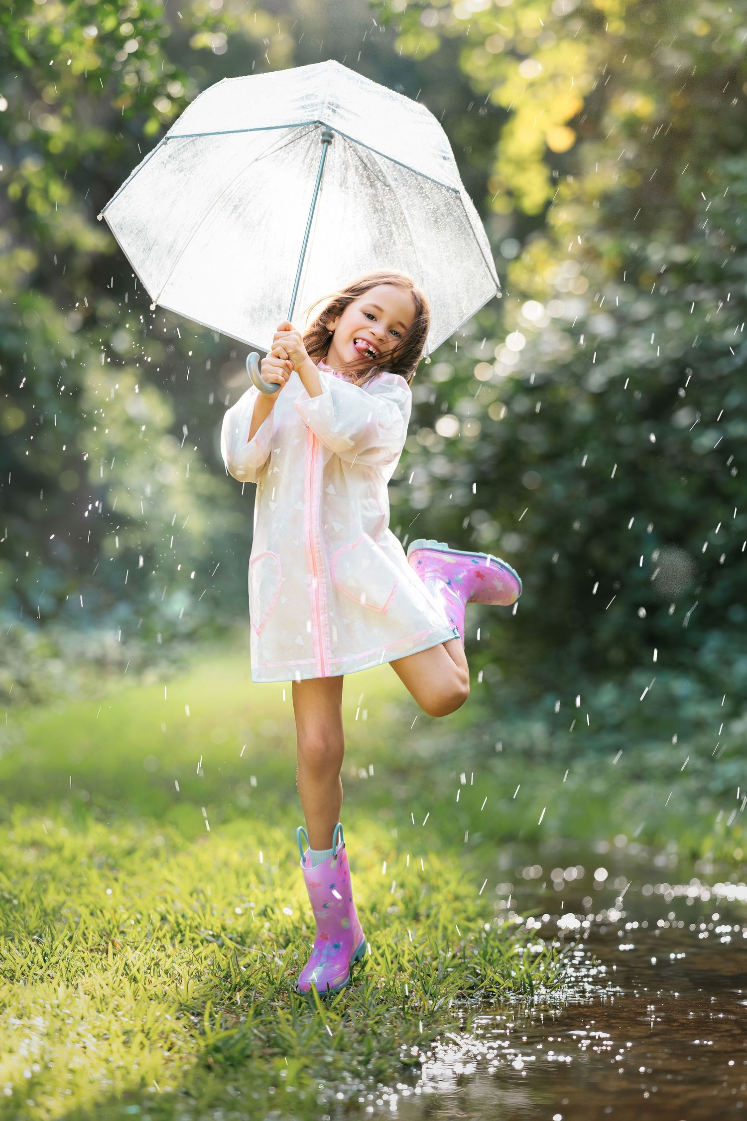 A joyful girl in a white dress and pink rain boots holds a clear umbrella while playing in the rain outdoors.