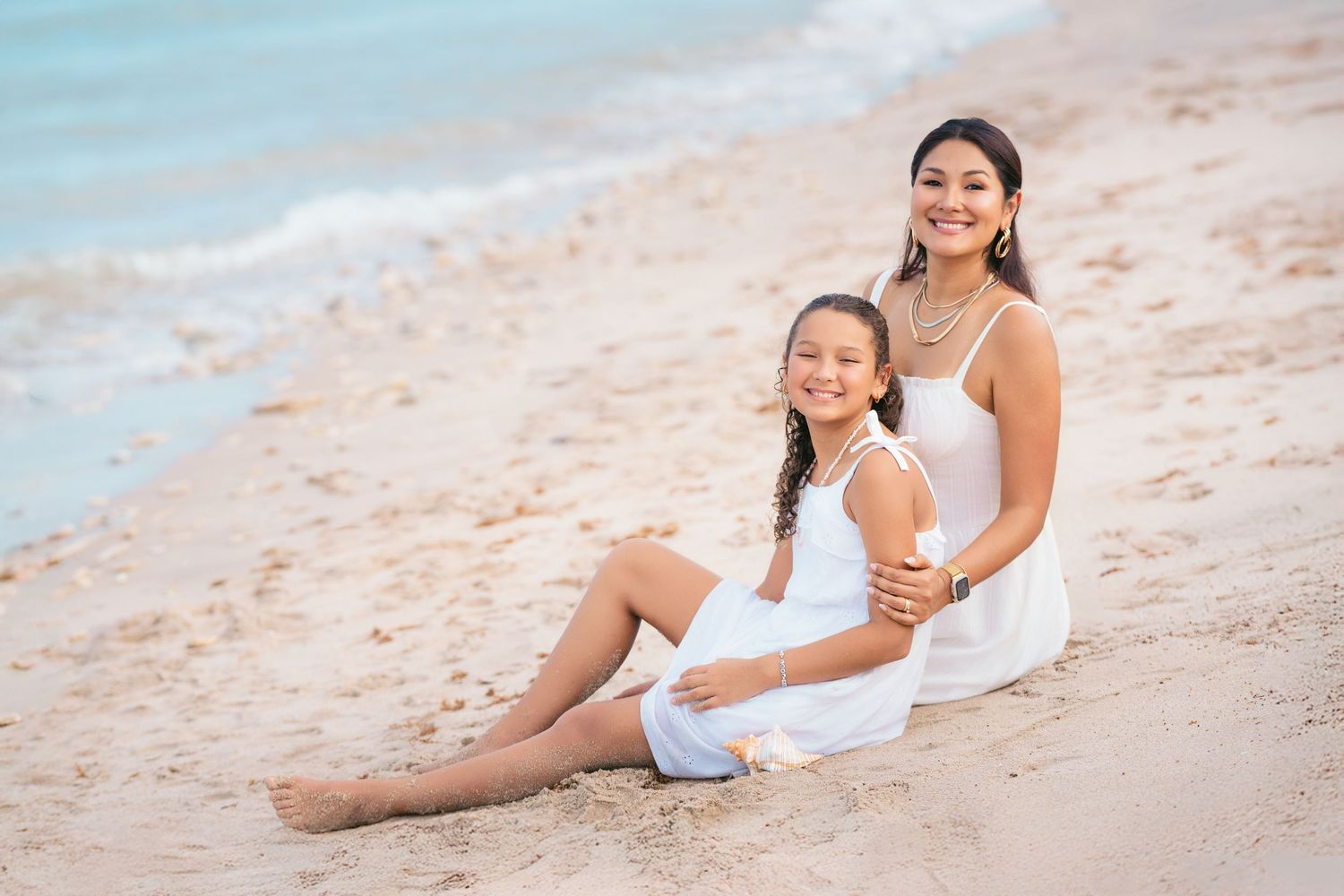 Woman and girl in white dresses sitting together on sandy beach with ocean waves in background.