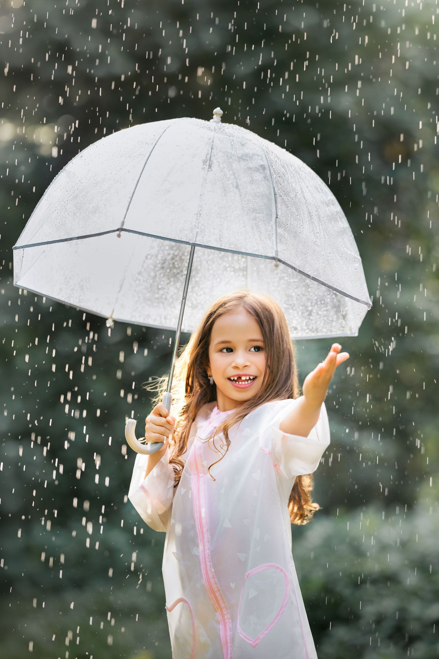 Happy young girl holding a clear umbrella in the rain, wearing a white raincoat, reaching out to catch raindrops.