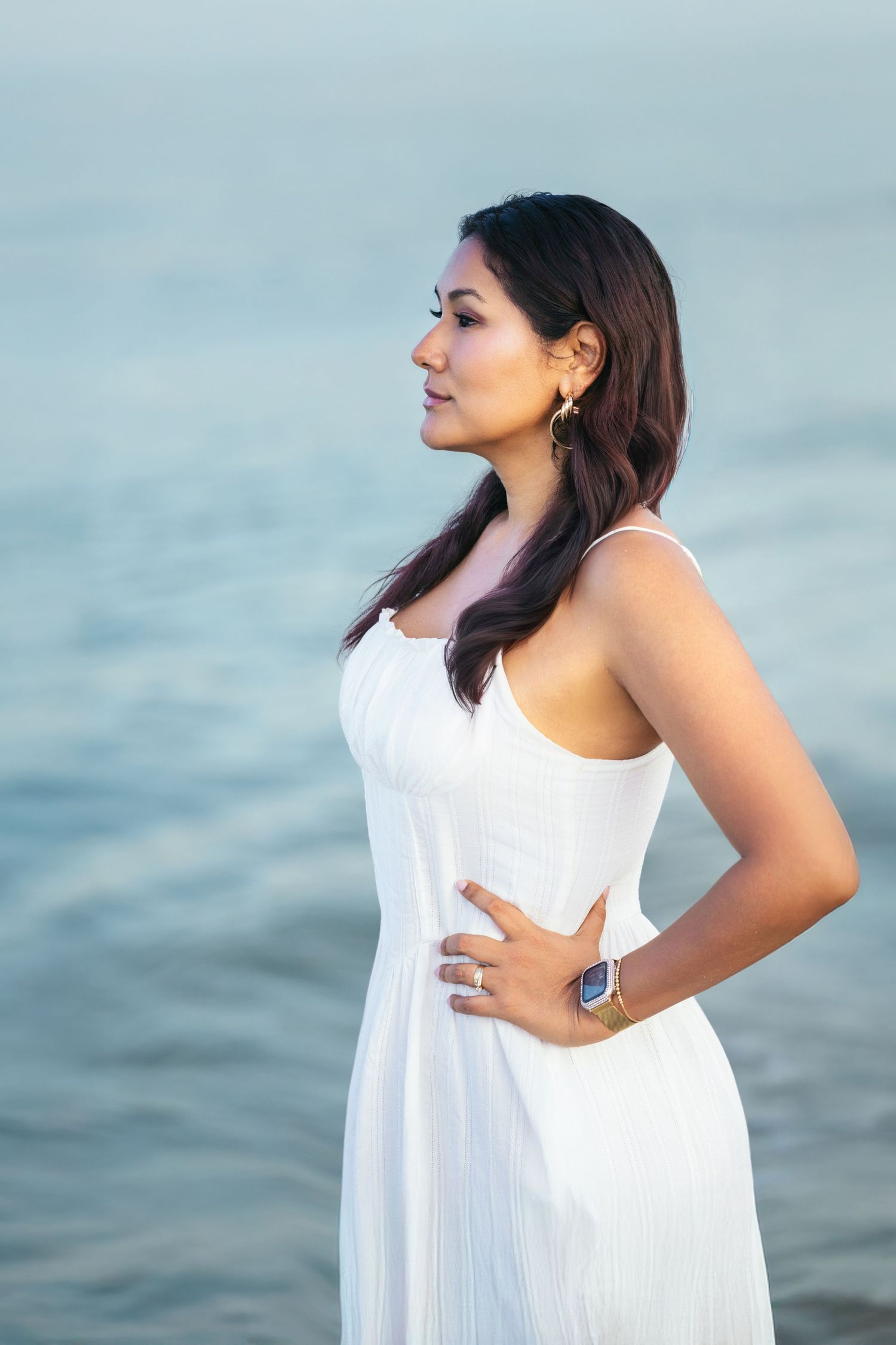Woman in white dress standing by the ocean, looking sideways with hand on hip, wearing gold earrings and a smartwatch.