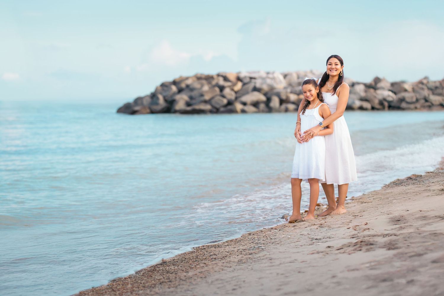 Mother and daughter in white dresses standing barefoot on a sandy beach with turquoise water and rocks.